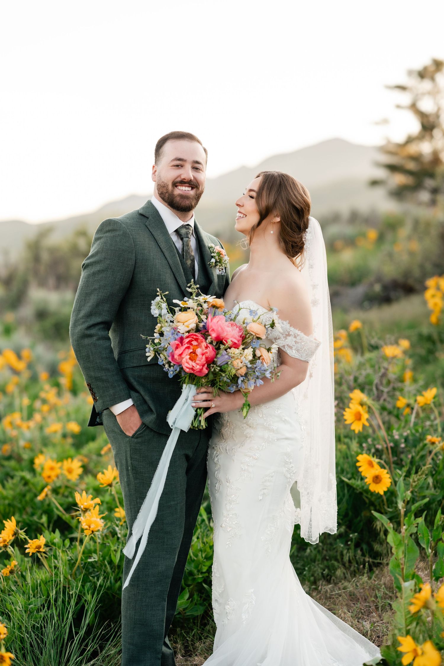 Newlyweds share joyful moments among blooming yellow wildflowers with mountains in the background.
