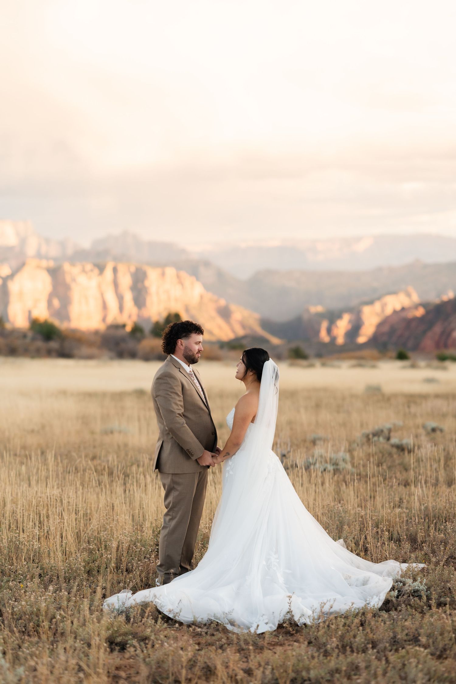 Newlyweds share an intimate moment in a field with mountains and sunset behind them.