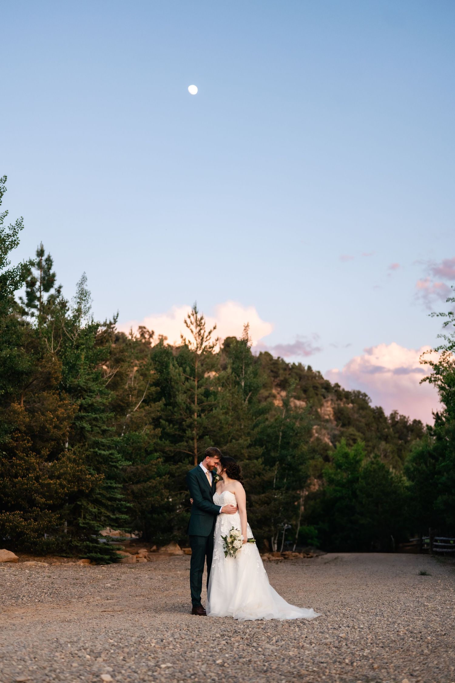 A couple shares an intimate moment at sunset in a mountain setting with pine trees and a full moon above.