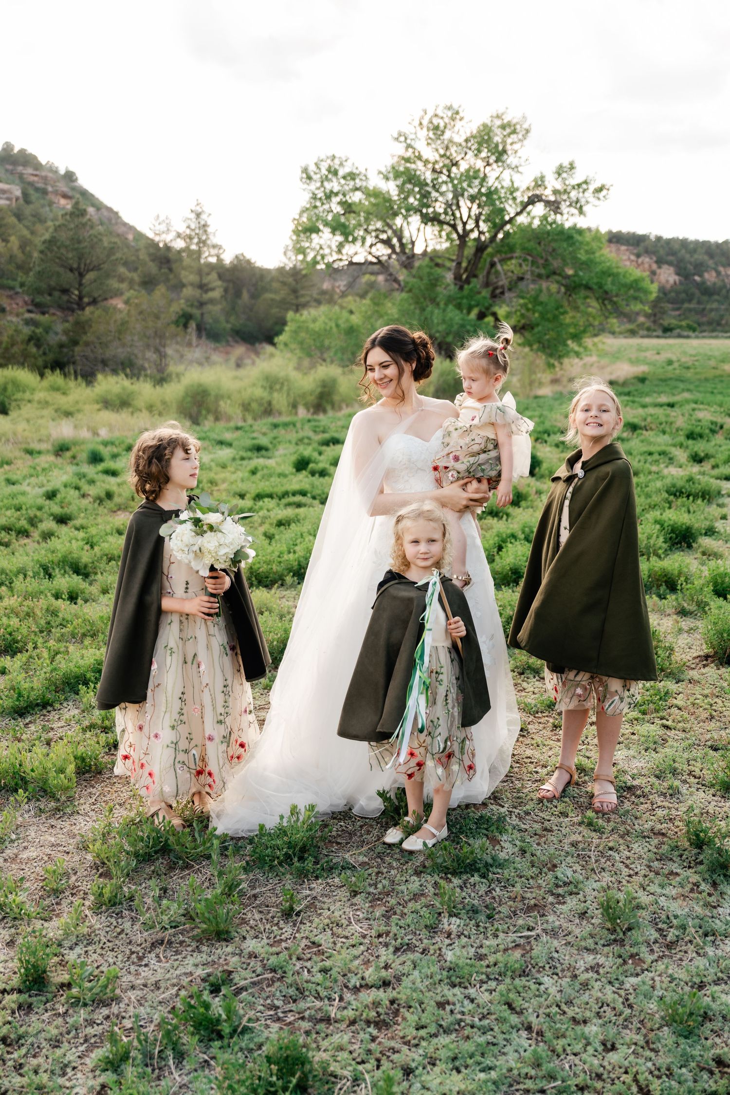 Bride in white gown poses with flower girls wearing floral dresses and dark coats in grassy field.