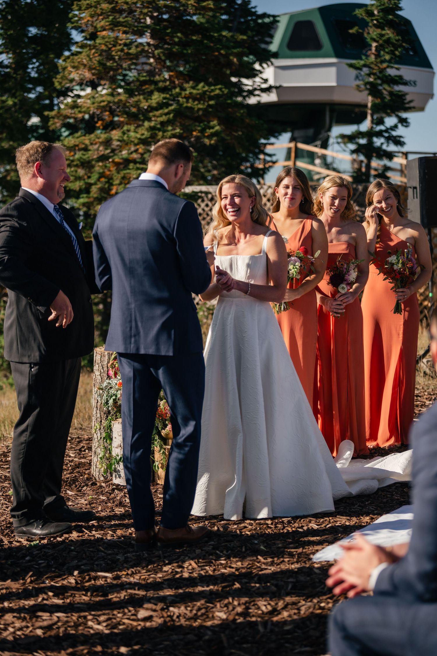 Wedding party gathers outdoors with bridesmaids wearing coral dresses against a natural mountain backdrop.