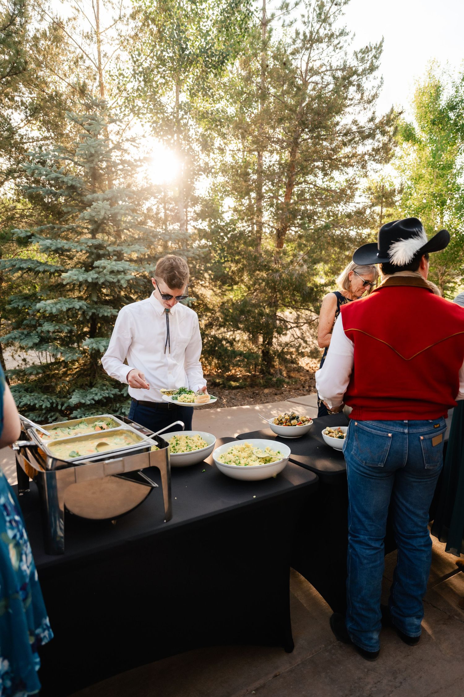 Buffet line setup outdoors with black tablecloths and food stations against a backdrop of evergreen trees.