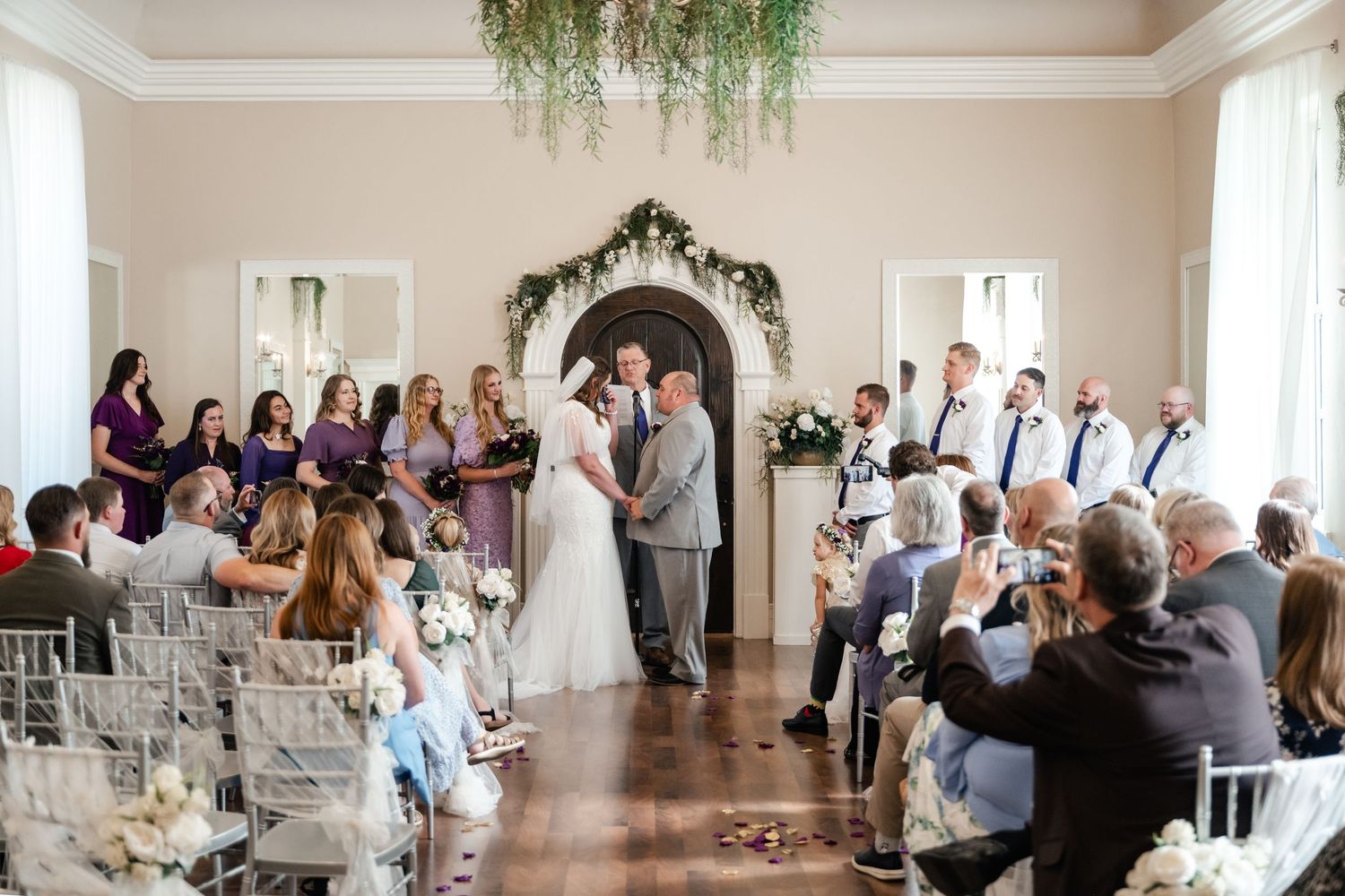 Wedding guests mingle and socialize in an elegant white ballroom with hanging greenery decorations during a reception. Stone Gate Venue.