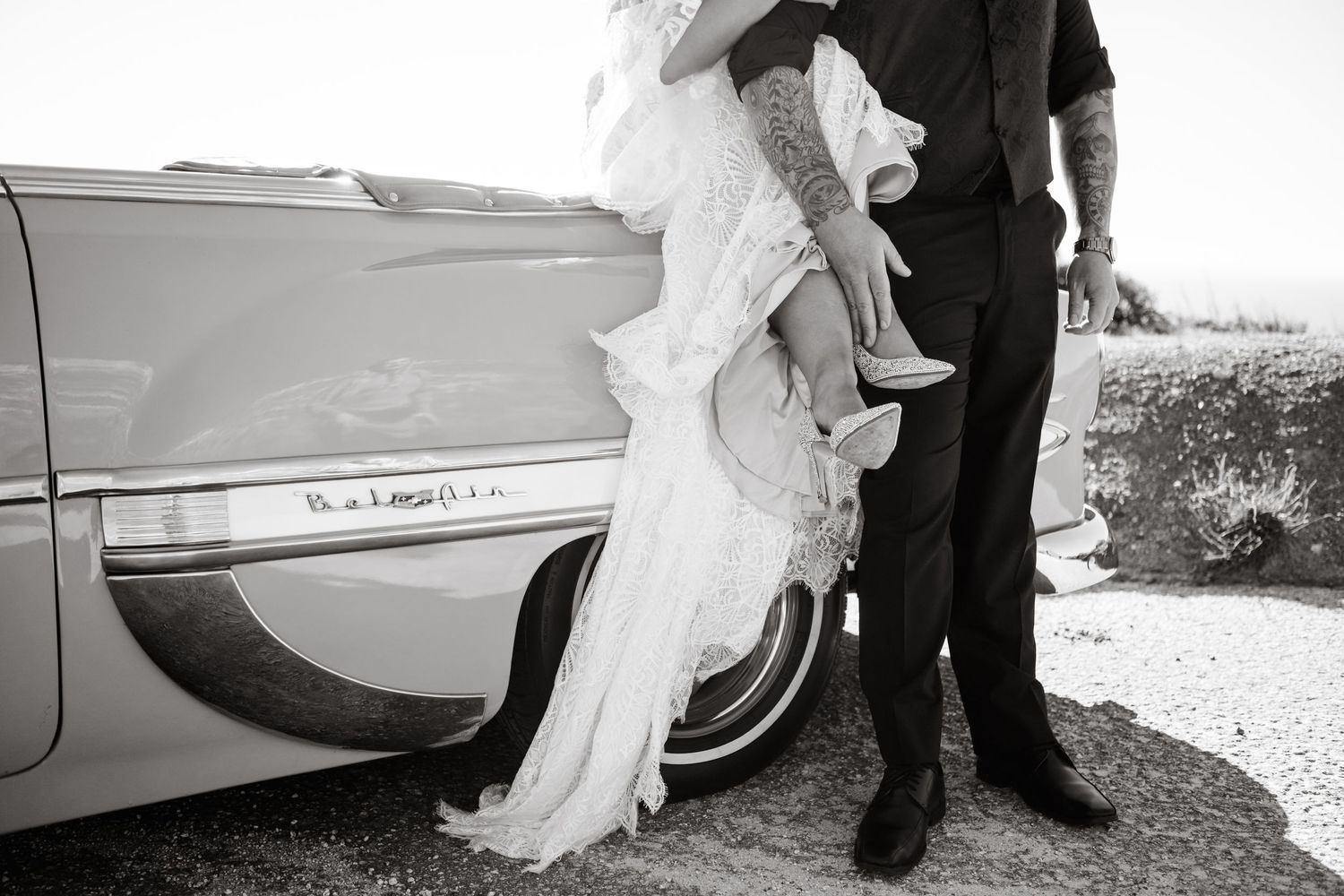 A black and white photo of a couple by a vintage car showing the bride's flowing lace dress.