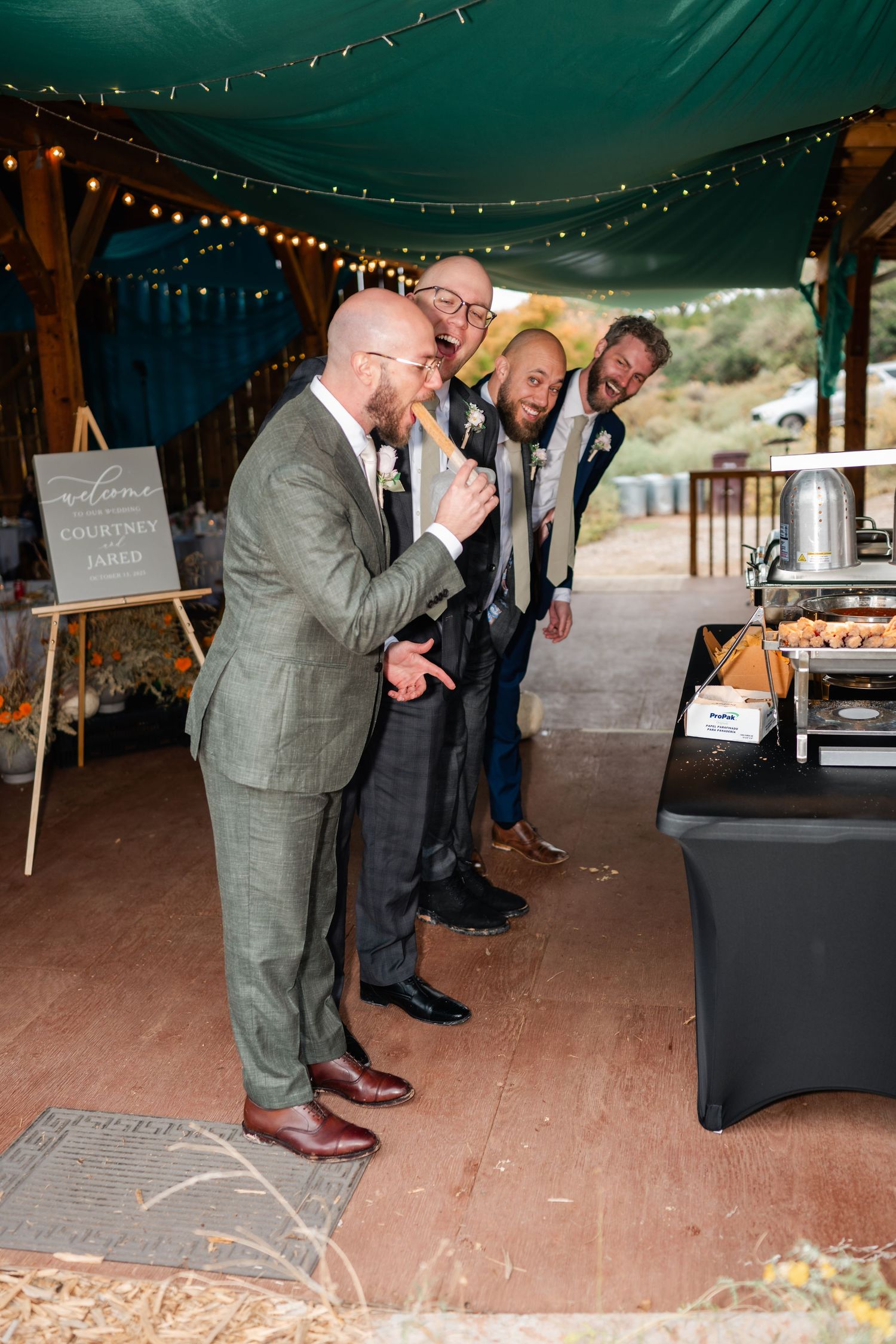 Wedding guests laugh and dance together on a covered wooden deck with string lights overhead.