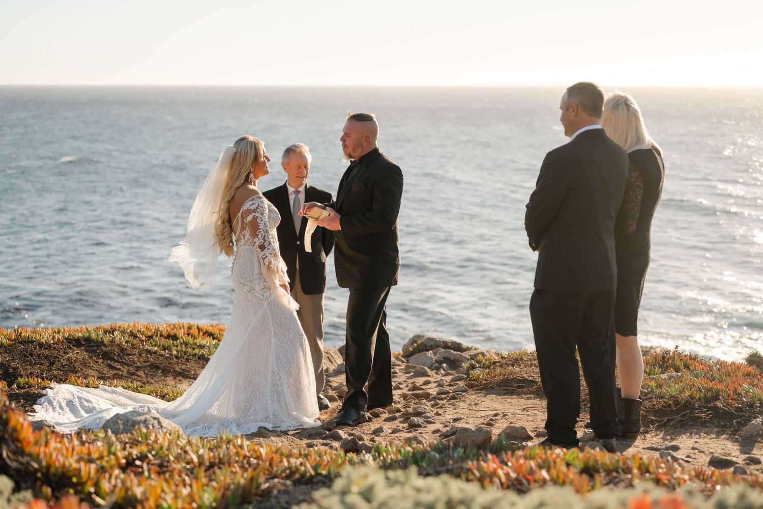 A wedding ceremony unfolds on a rugged coastline with golden light illuminating the scene.
