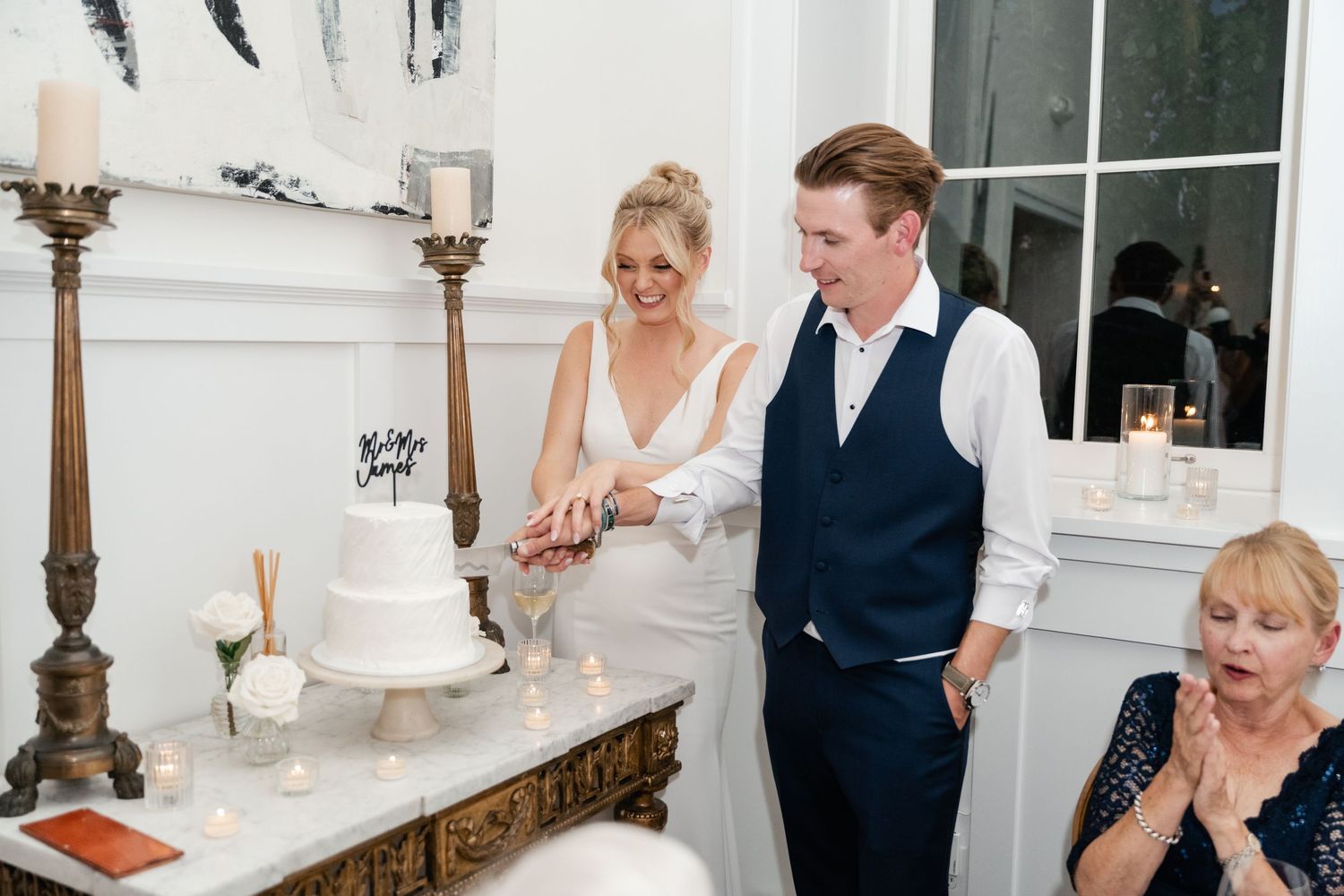Bride and groom cutting white wedding cake at elegant indoor reception with candles and floral decor.