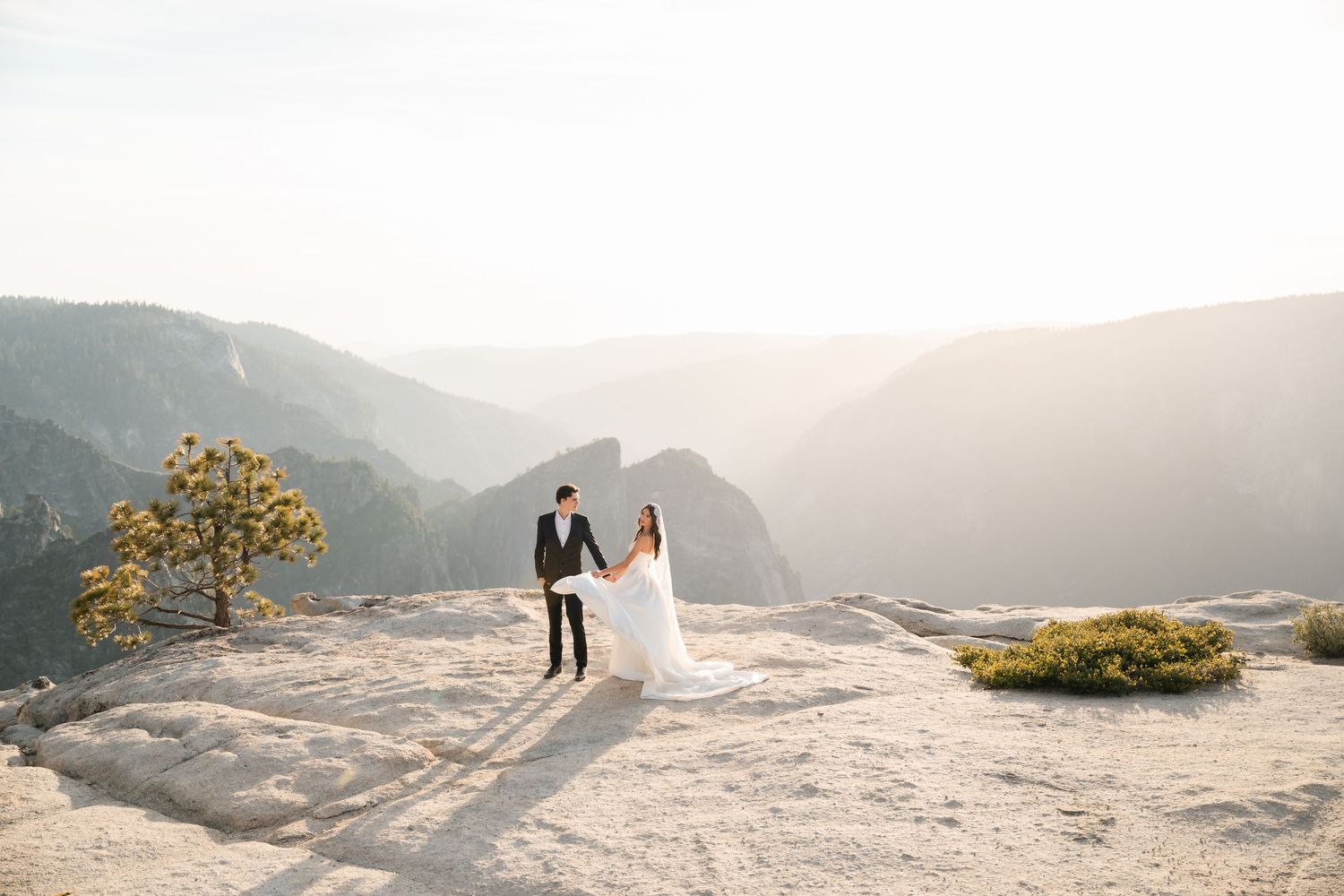 Bride and groom hold hands on a rocky mountain overlook at Yosemite, with misty valley views behind them.