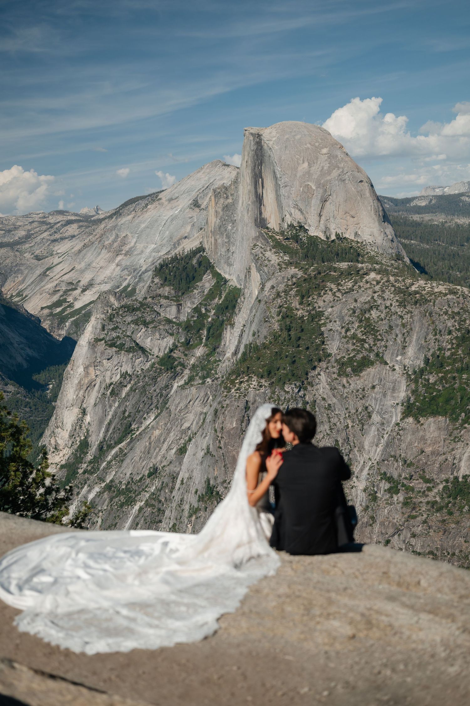 Bride and groom sit on Glacier Point rock ledge with Half Dome mountain backdrop at Yosemite National Park.