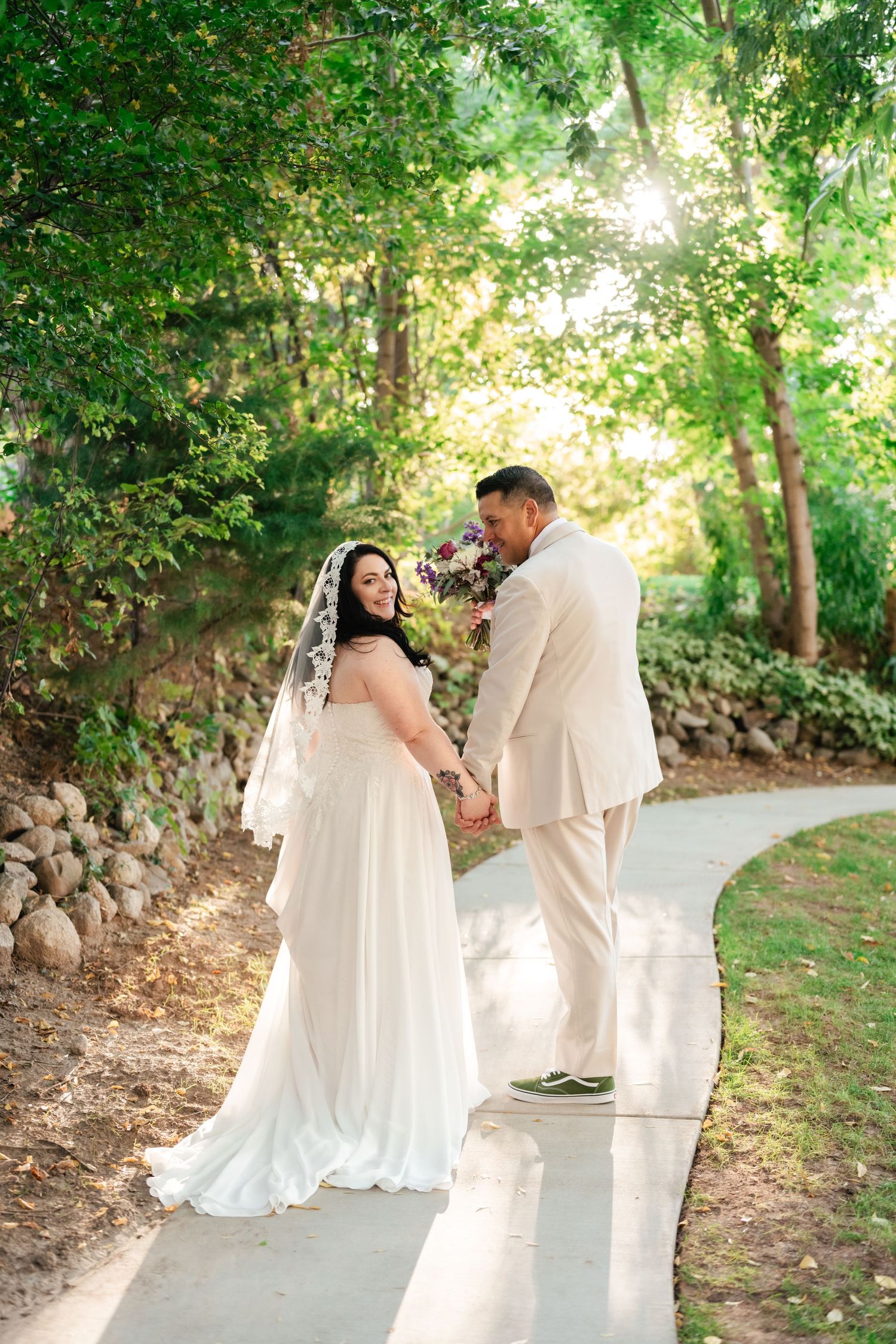 A romantic wedding couple poses on a sunlit garden path while wearing elegant white and cream attire.
