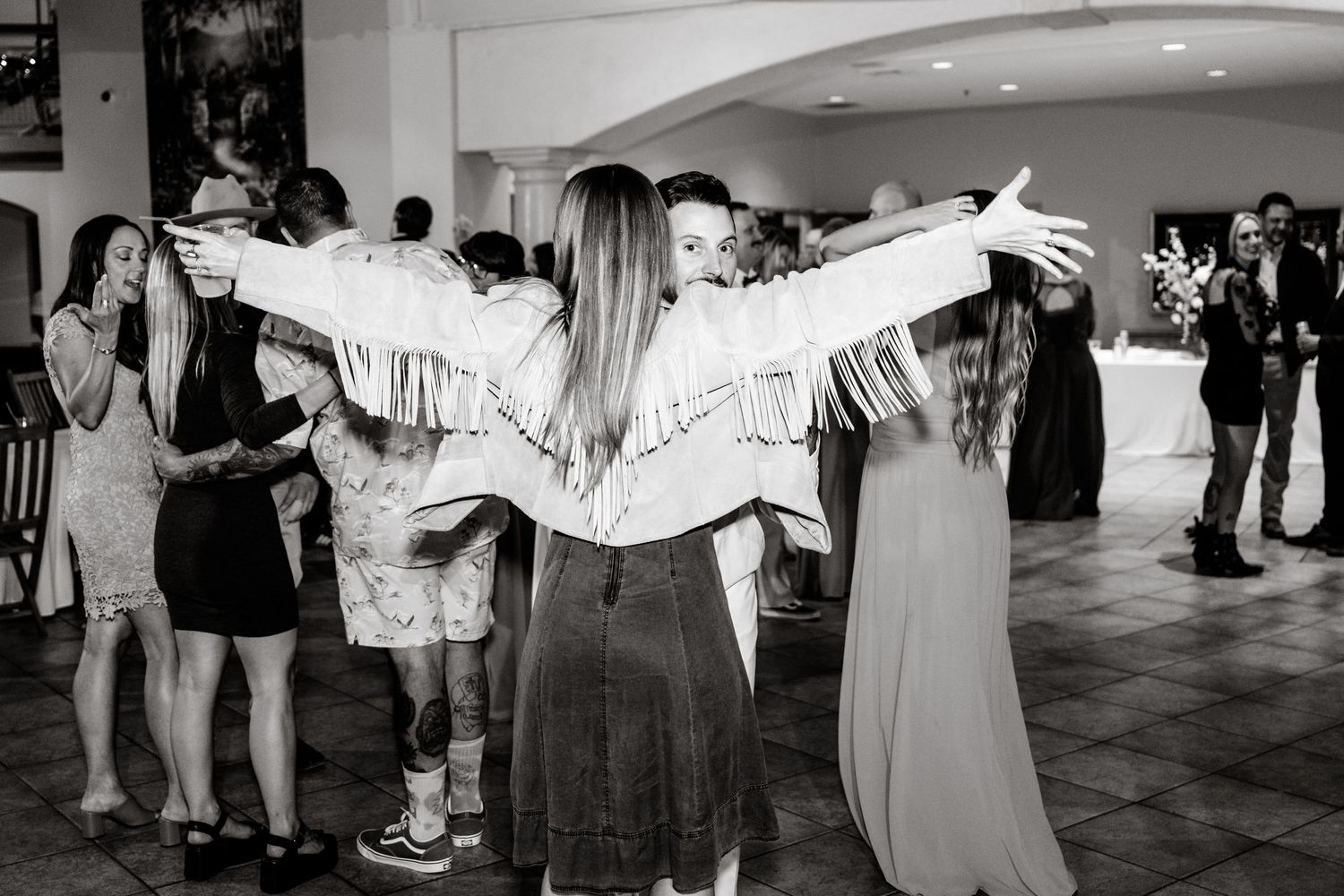 A person wearing a fringed denim jacket with arms outstretched dances at a party among other guests in monochrome.