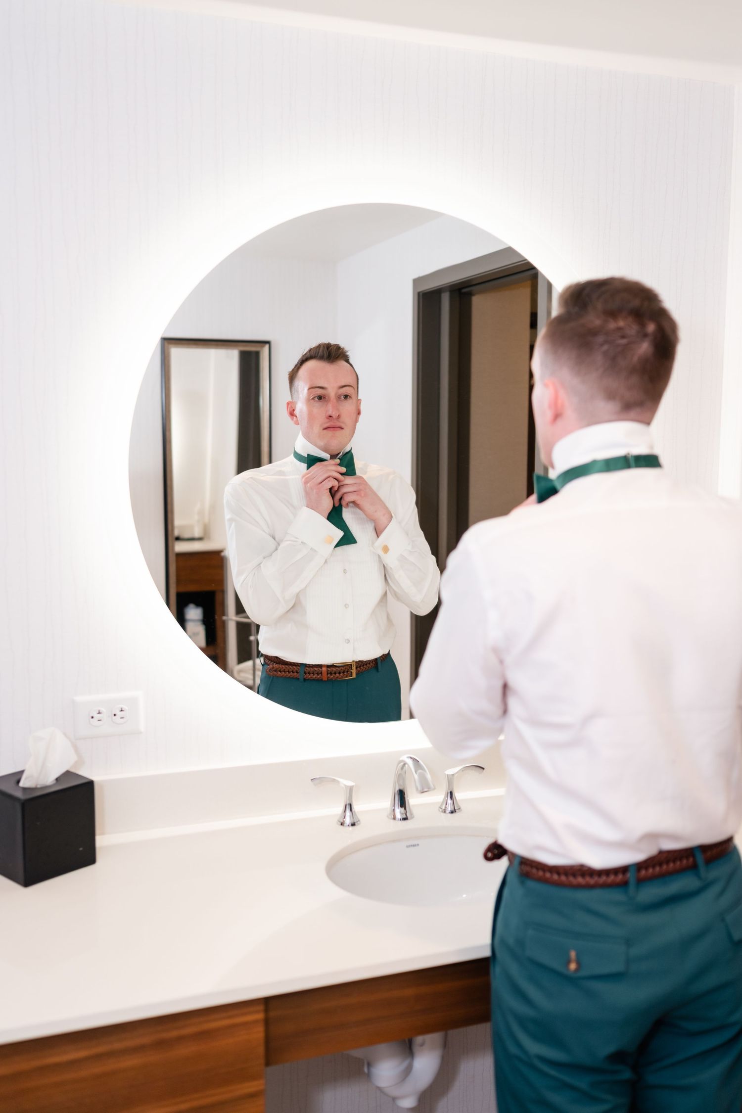 A person in a white dress shirt and green pants adjusts their bowtie while looking in a bathroom mirror.