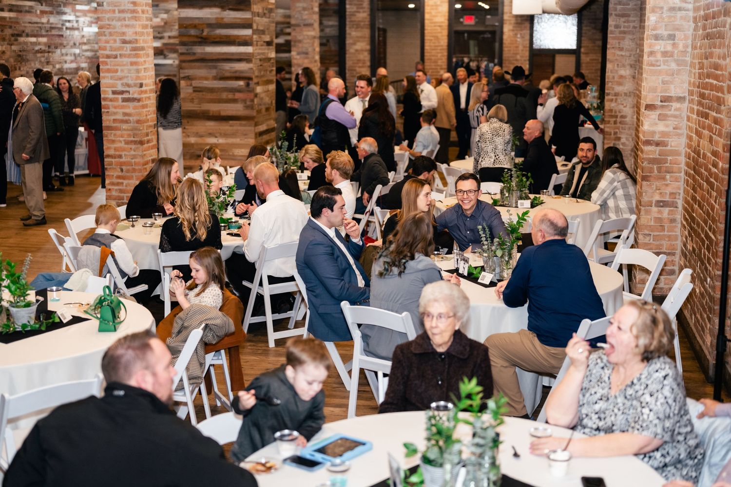 Reception dinner with guests seated at tables in rustic brick venue with white chairs and greenery centerpieces.