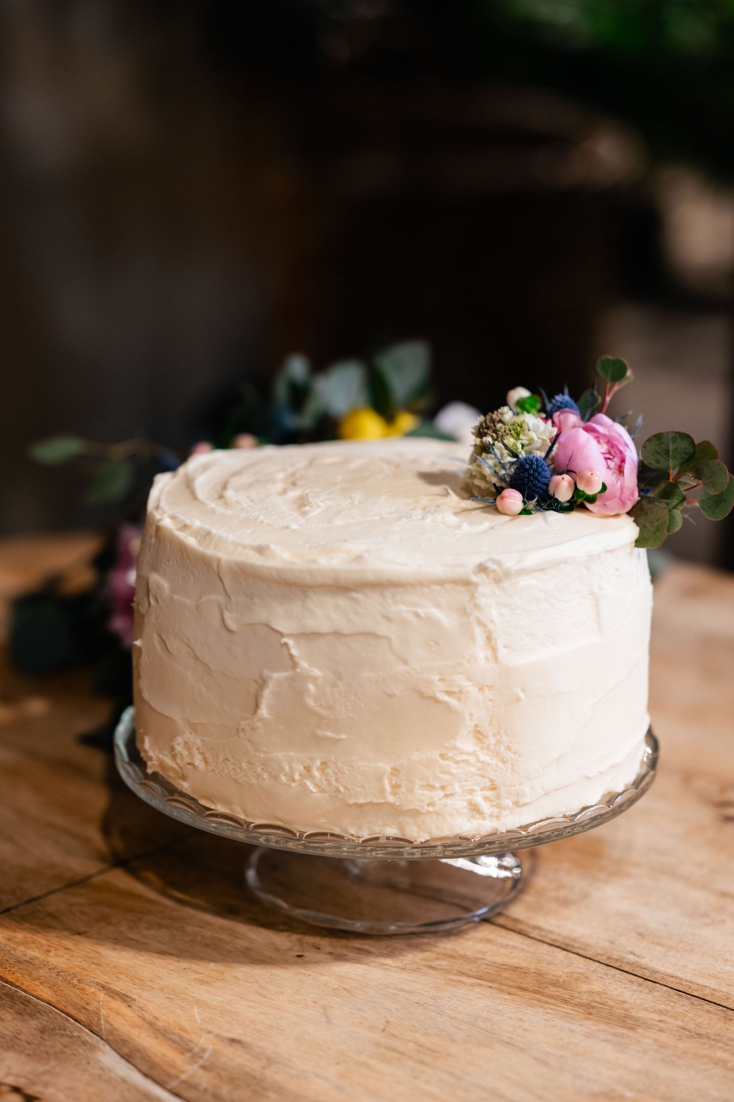 Elegant white frosted cake decorated with fresh flowers sits on a glass cake stand atop wooden table.