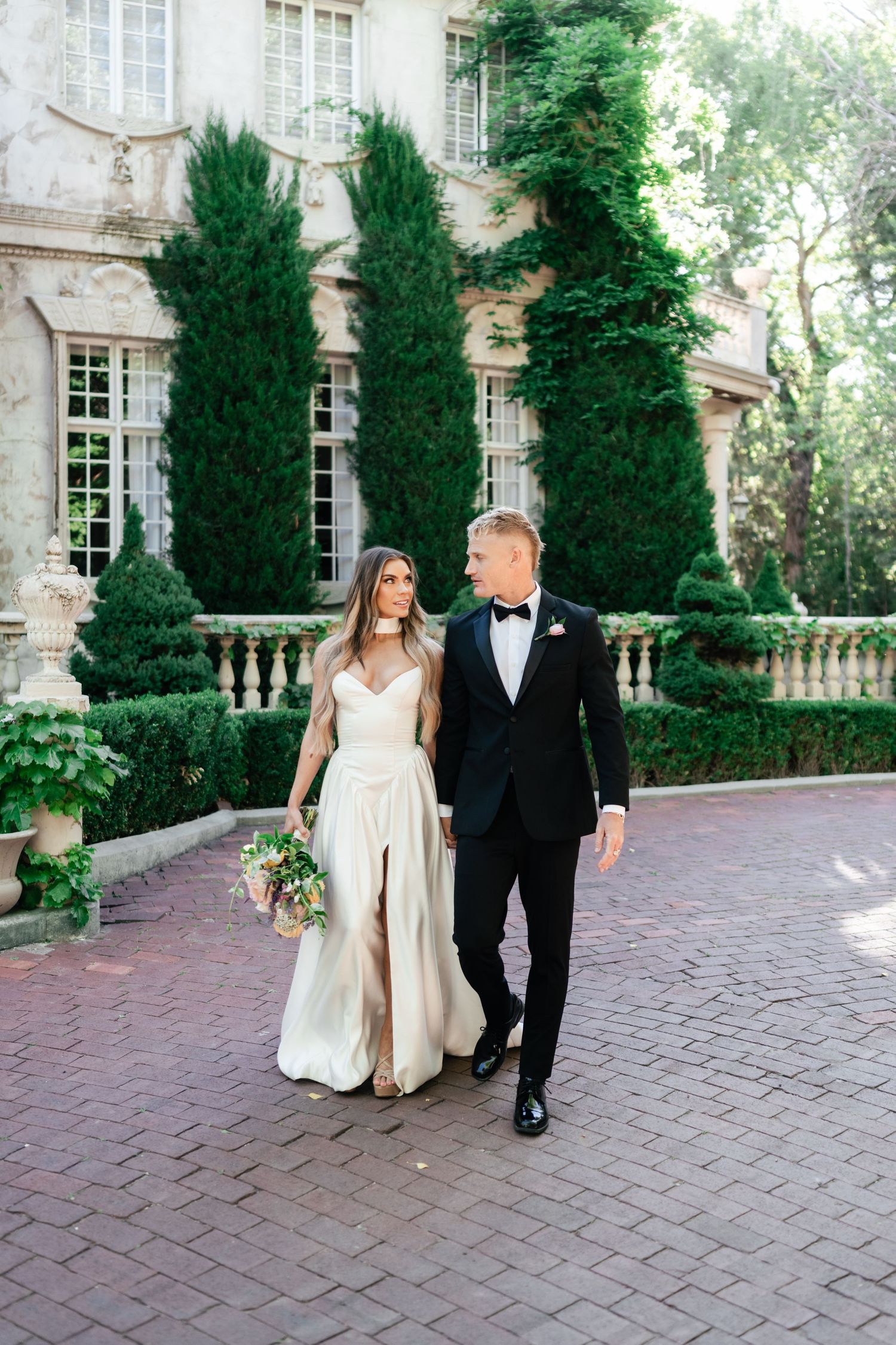 A couple in formal wedding attire walking and posing for portraits along a brick walkway at an elegant stone mansion.