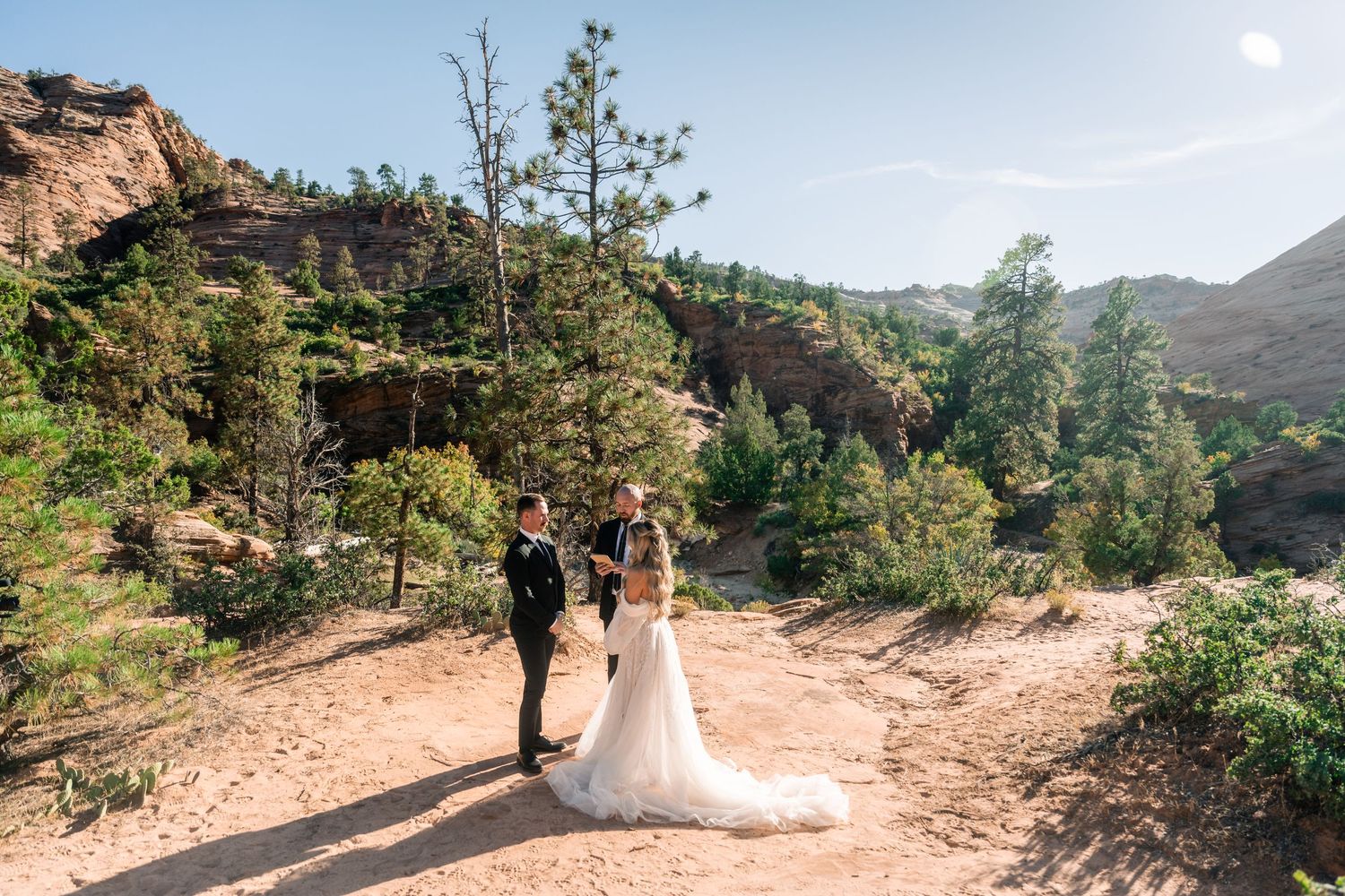 A wedding couple walks along a desert path with dramatic red rock cliffs in the background.