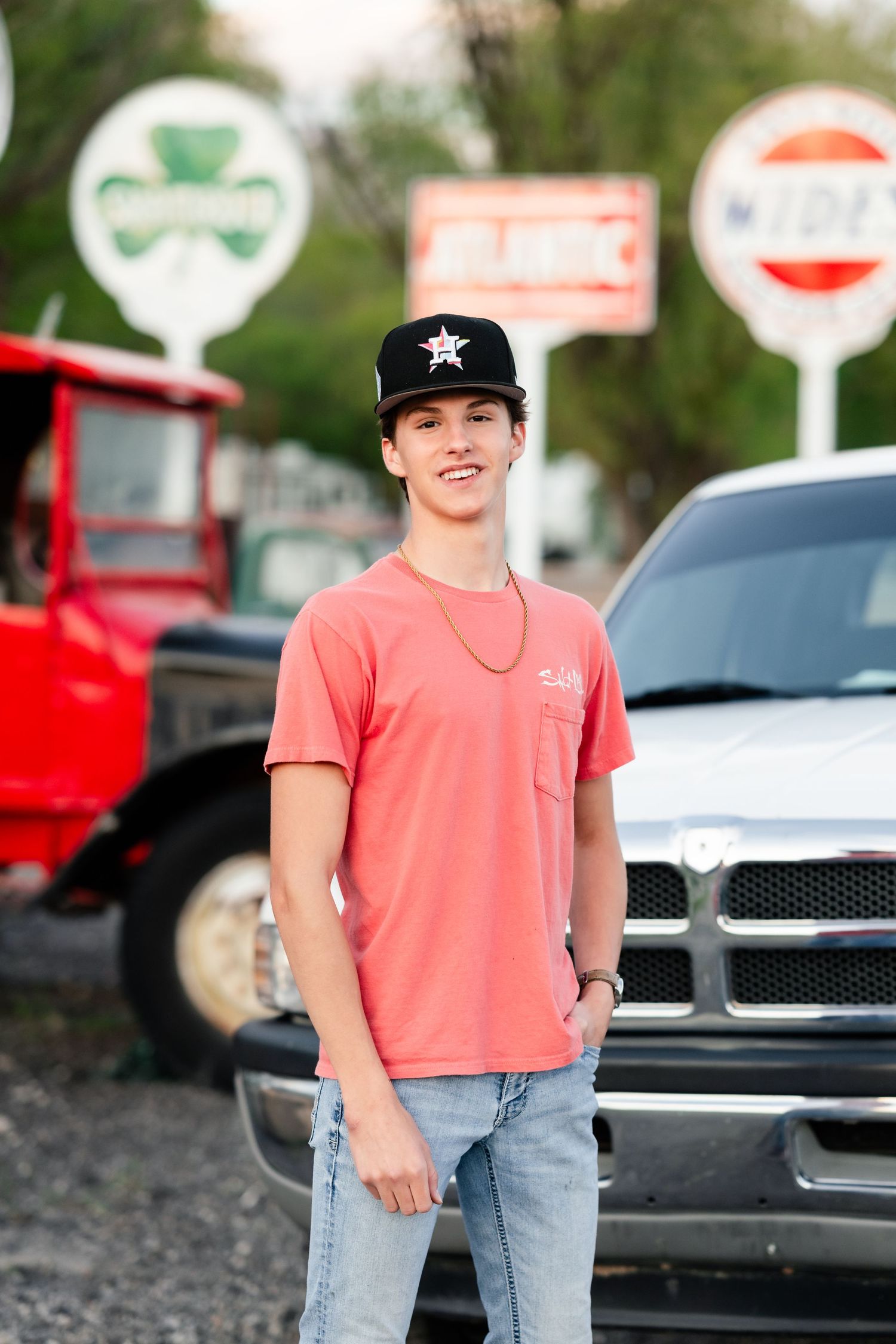 Person wearing a coral t-shirt and baseball cap poses near parked cars and traffic signs.