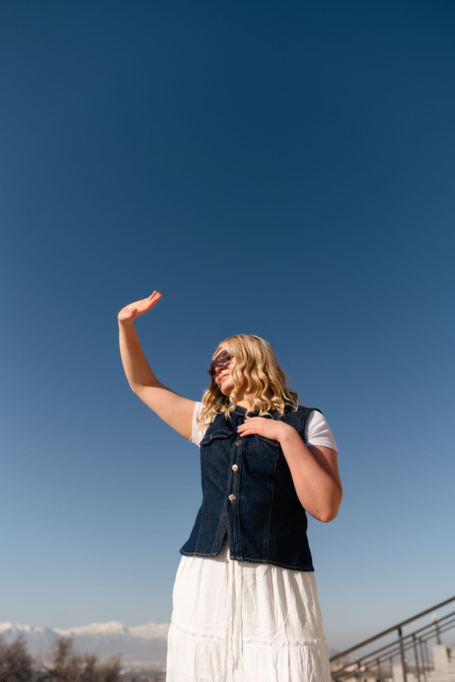 Woman in denim vest and white dress poses against blue sky with raised arm and sunglasses on sunny day.