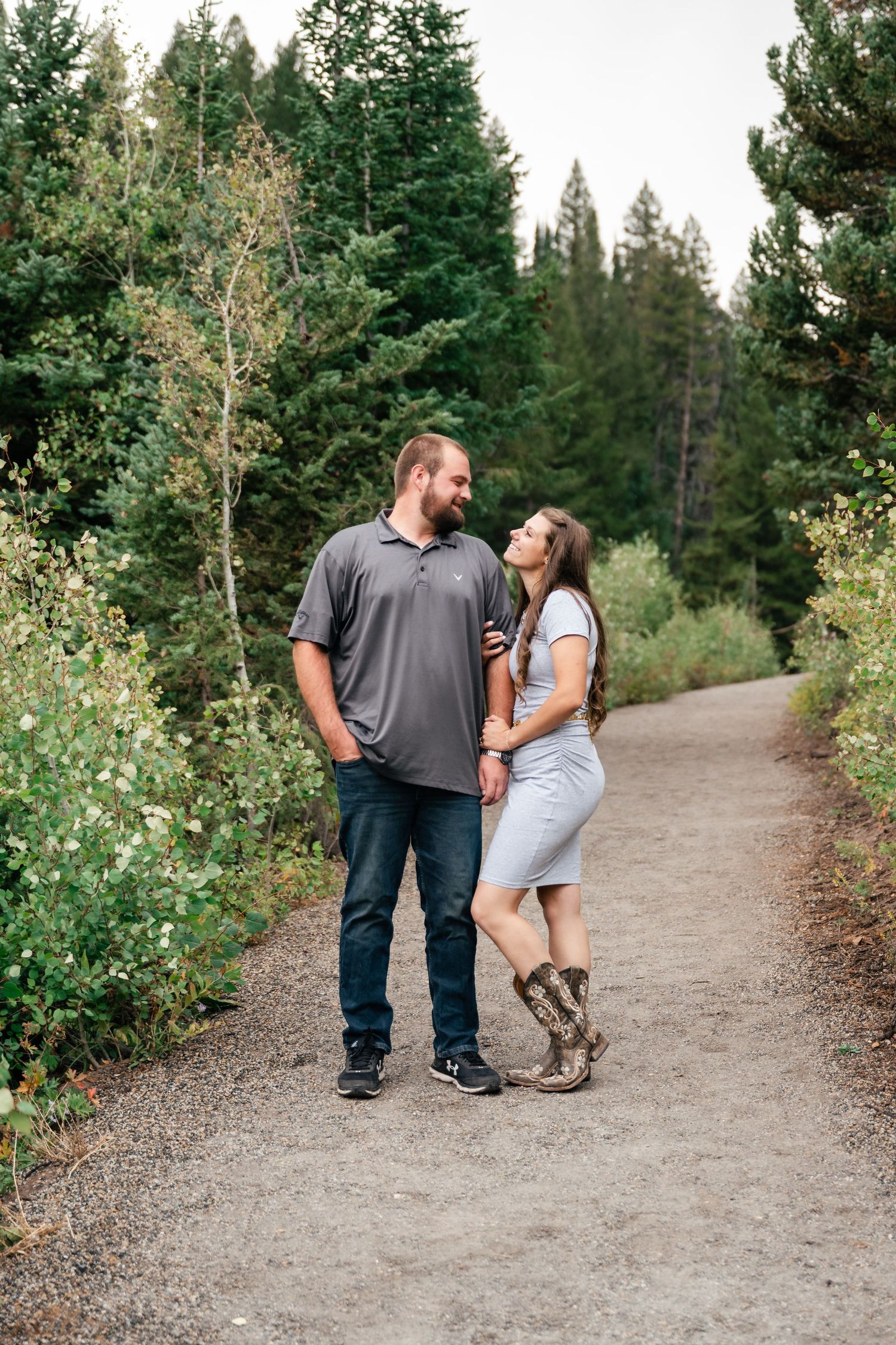 A couple shares a romantic moment while standing on a forest trail surrounded by evergreen trees and aspen foliage.
