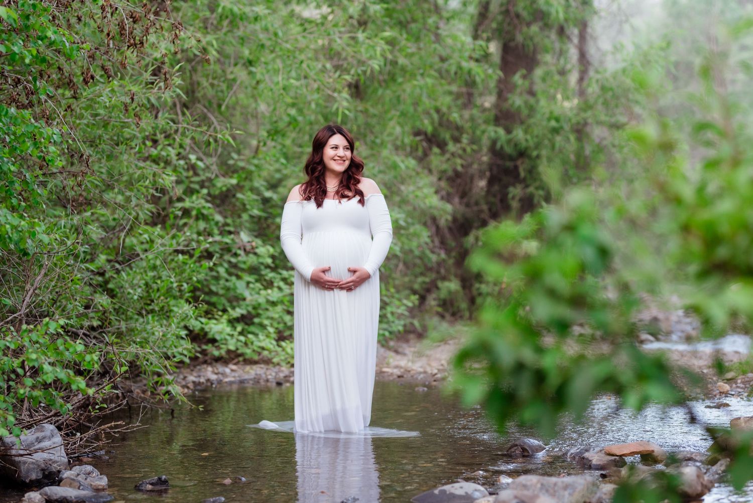 A figure in a white dress stands gracefully by a flowing stream in a lush green forest setting.