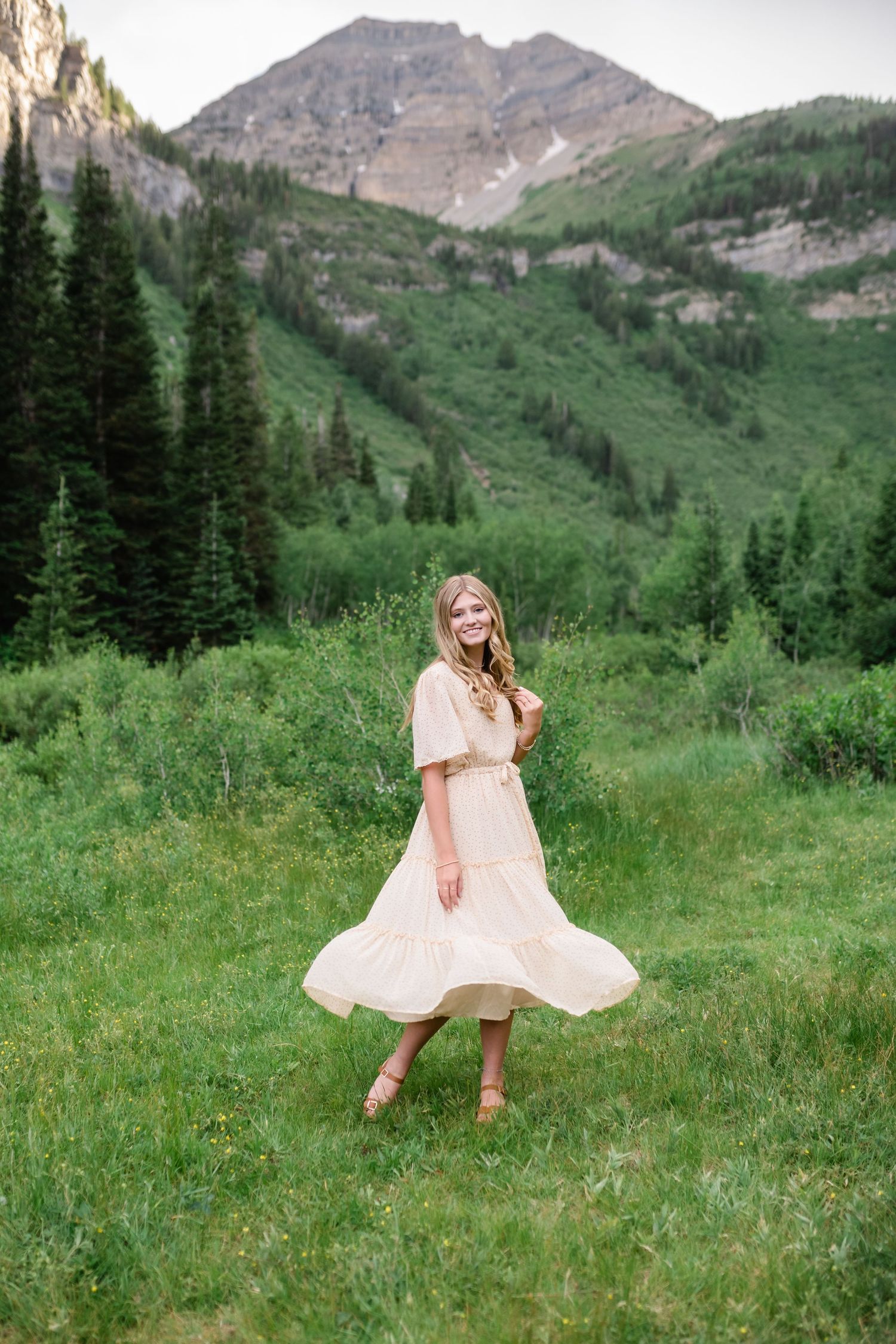 A person in a flowing peach dress twirls playfully in a green mountain meadow with majestic peaks in the background.