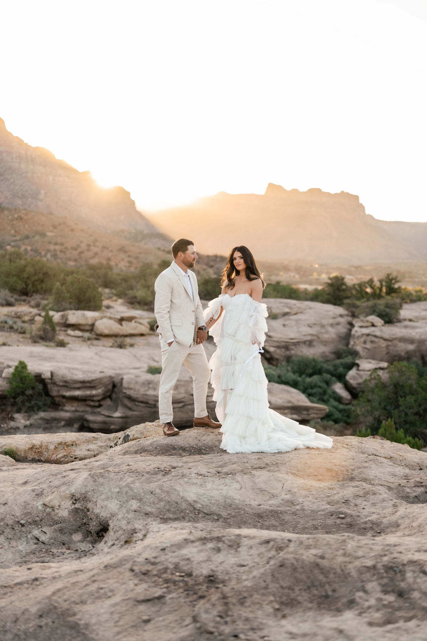 Zion destination photographer.  A couple in white attire shares an intimate moment on rocky terrain during golden hour sunset.