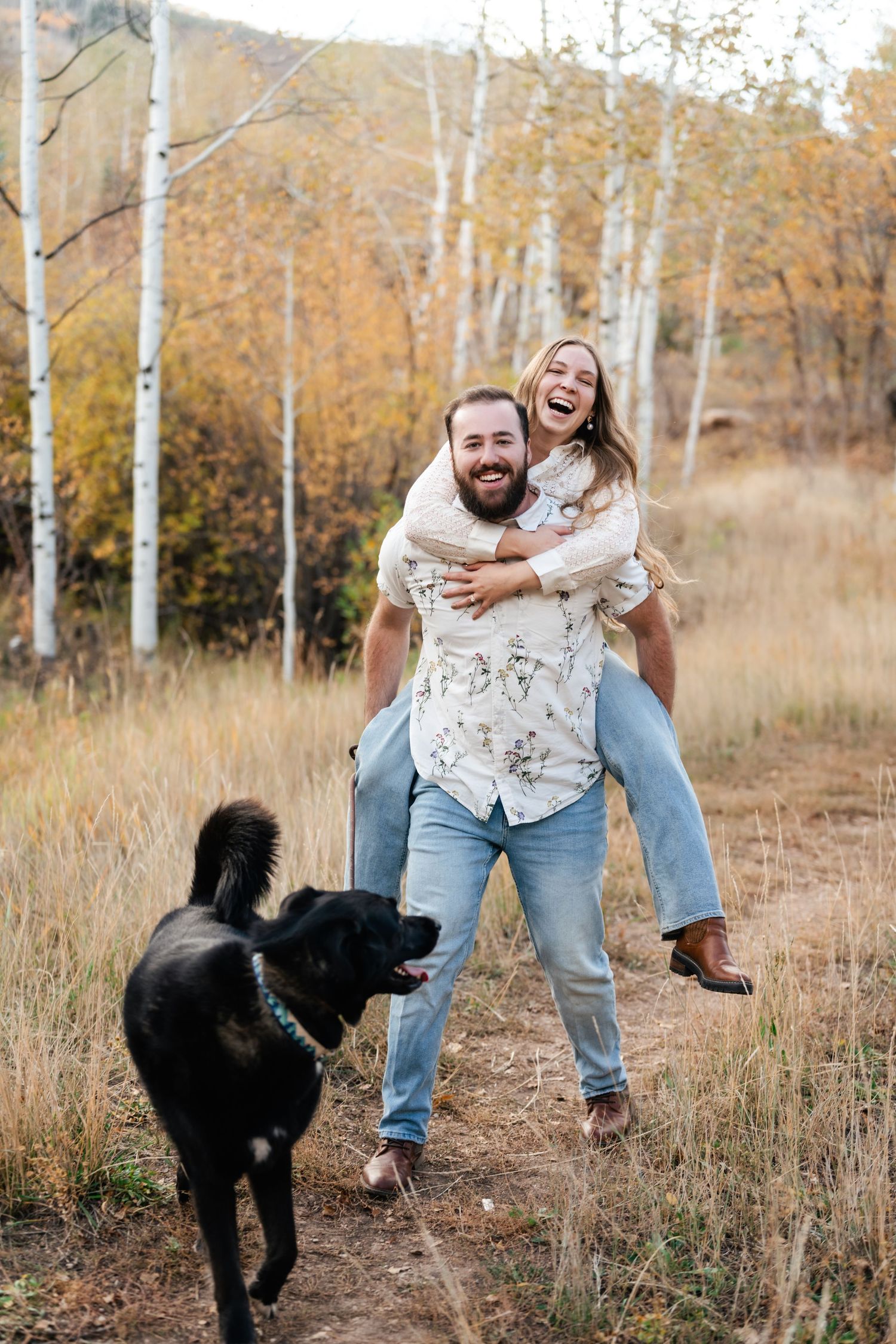 Playful couple and their black dog enjoying a fun photoshoot in an autumn field with birch trees. Park City Engagement Photographer.