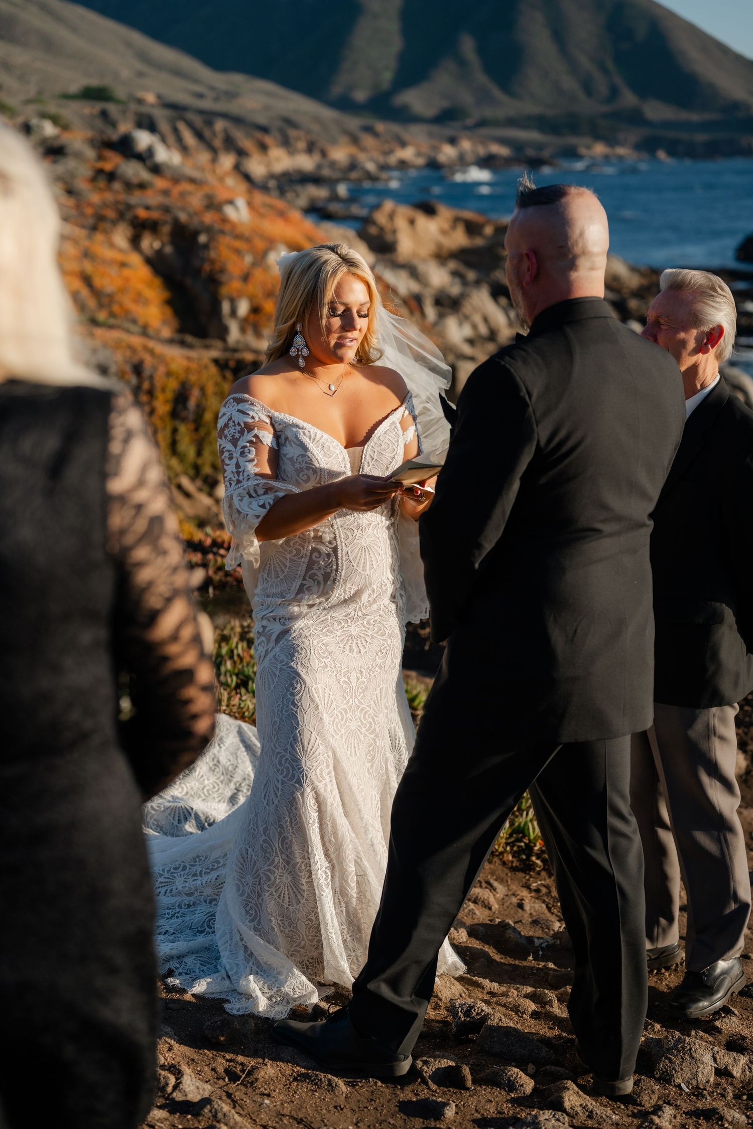 Wedding ceremony on rugged coastal cliffs with dramatic mountain backdrop and ocean views.