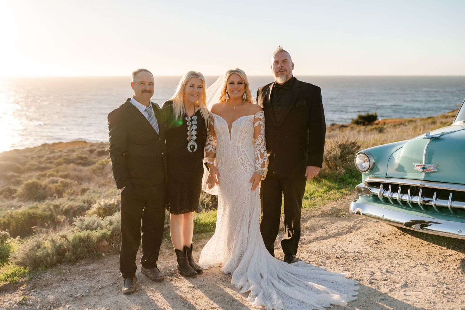 A wedding party poses together at sunset on a coastal cliff with a vintage turquoise car in the background.