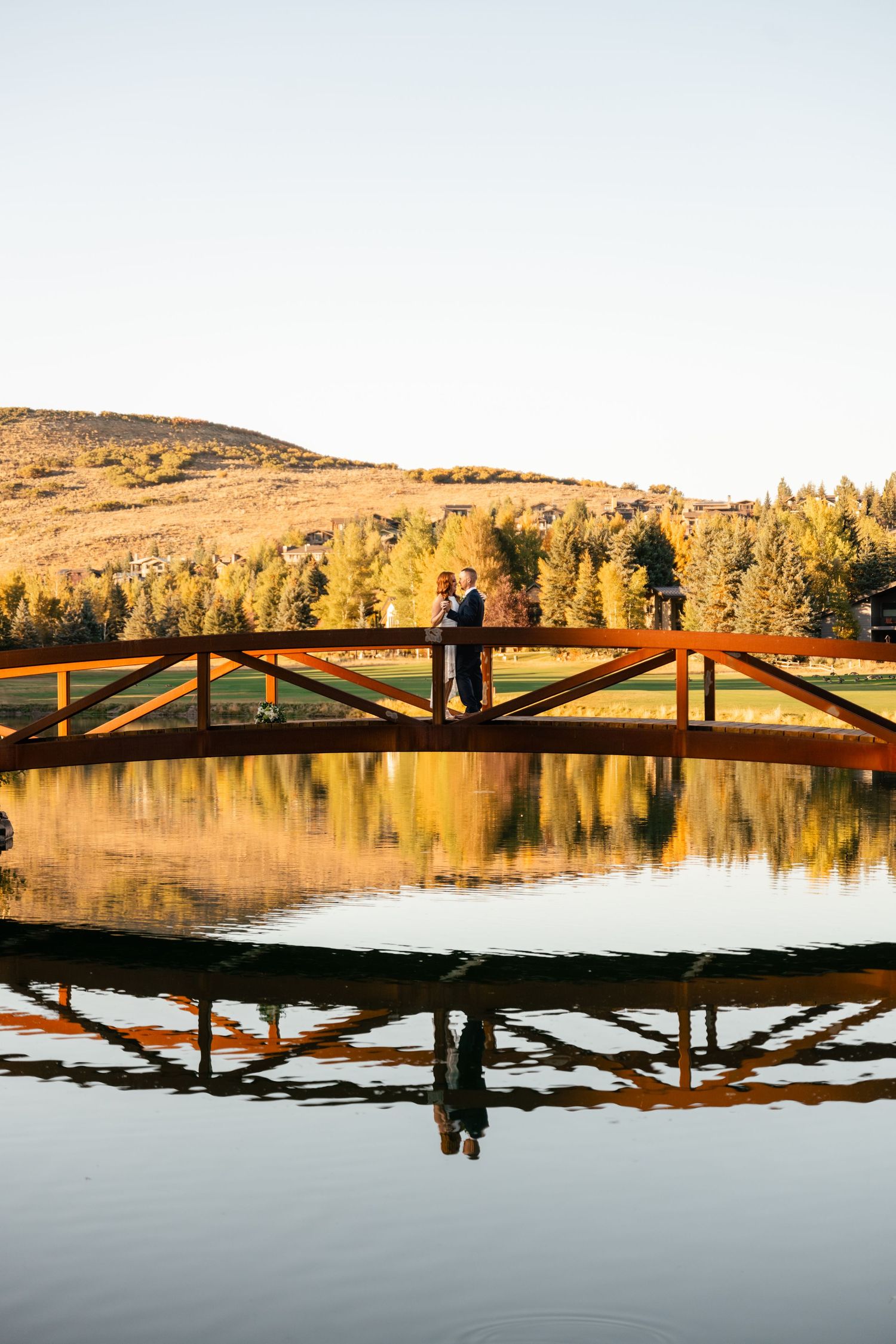 Silhouettes of figures stand on a wooden bridge at sunset with golden reflections on a tranquil lake.