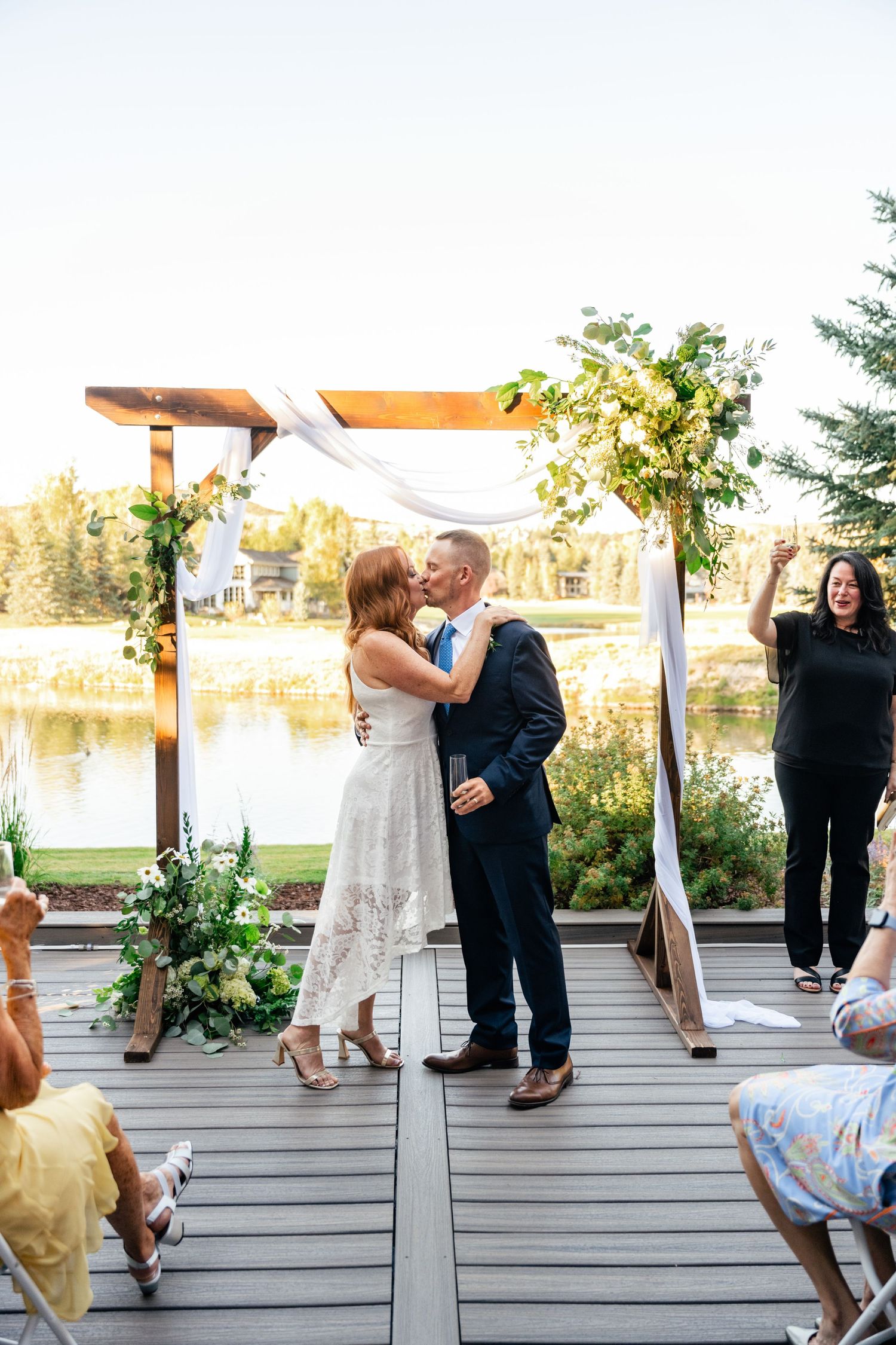 Casual outdoor wedding ceremony with guests seated on a wooden deck overlooking a lake.