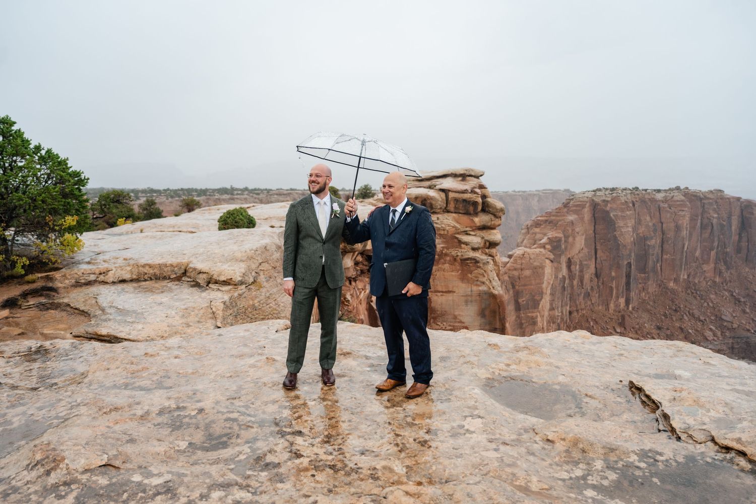 Three people standing on rocky cliff edge with canyon vista in background.