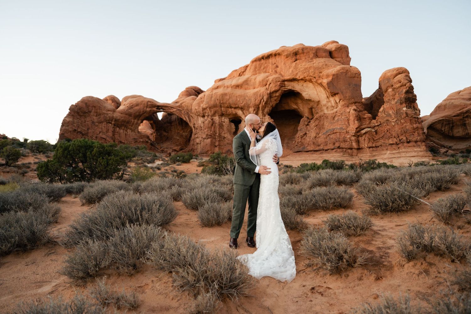 A couple shares their wedding moment against a backdrop of natural desert arches at sunset.