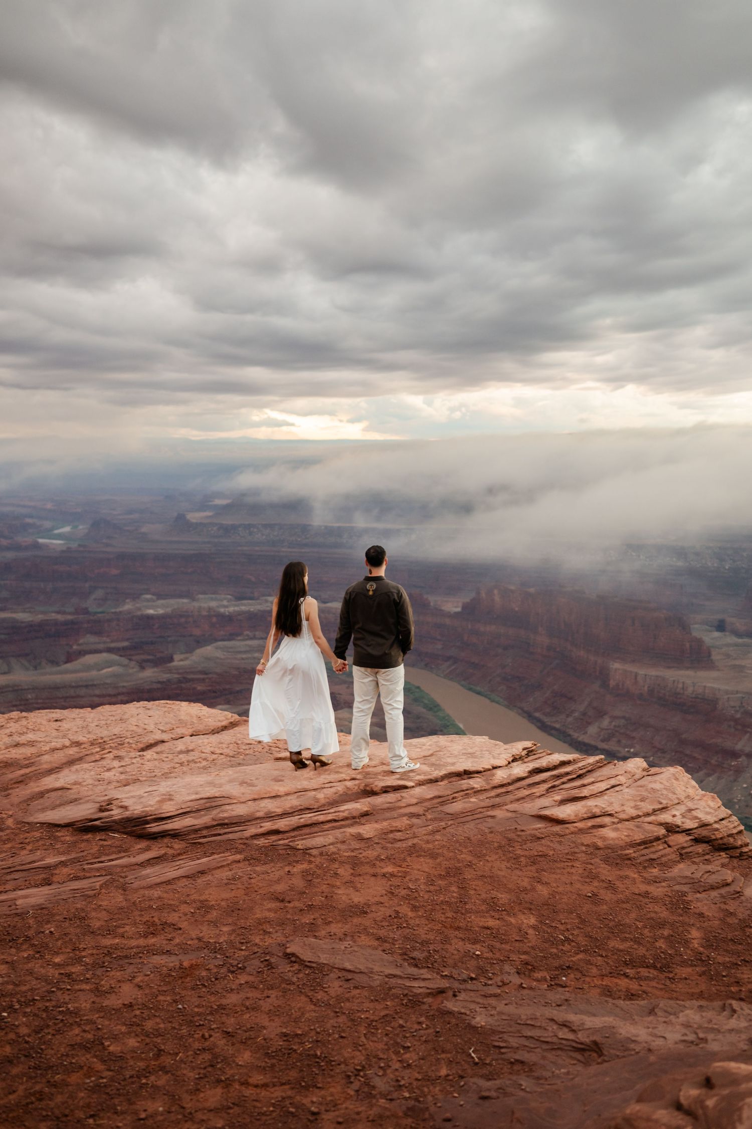 Couple holding hands at Dead Horse Point overlook, gazing at dramatic canyon views under a cloudy sky.