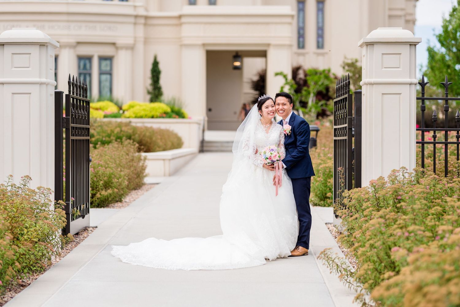Bride and groom pose together on a garden pathway outside an elegant wedding venue with lush greenery.