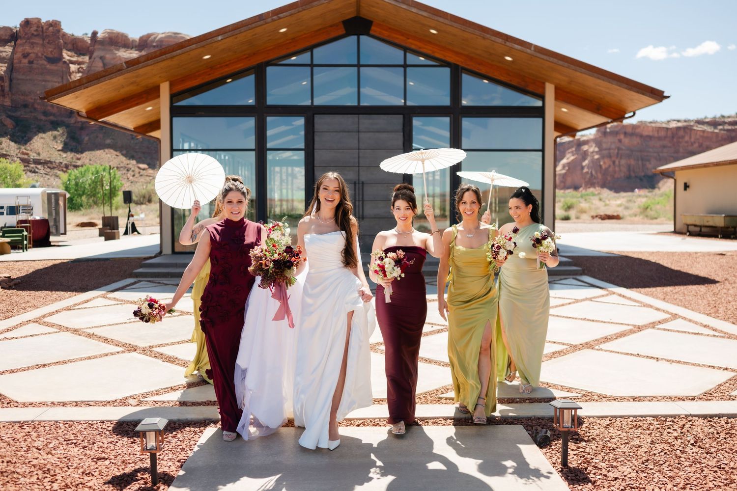 Bride and four bridesmaids in burgundy and green dresses holding parasols and bouquets outside a modern desert wedding venue in Moab, utah