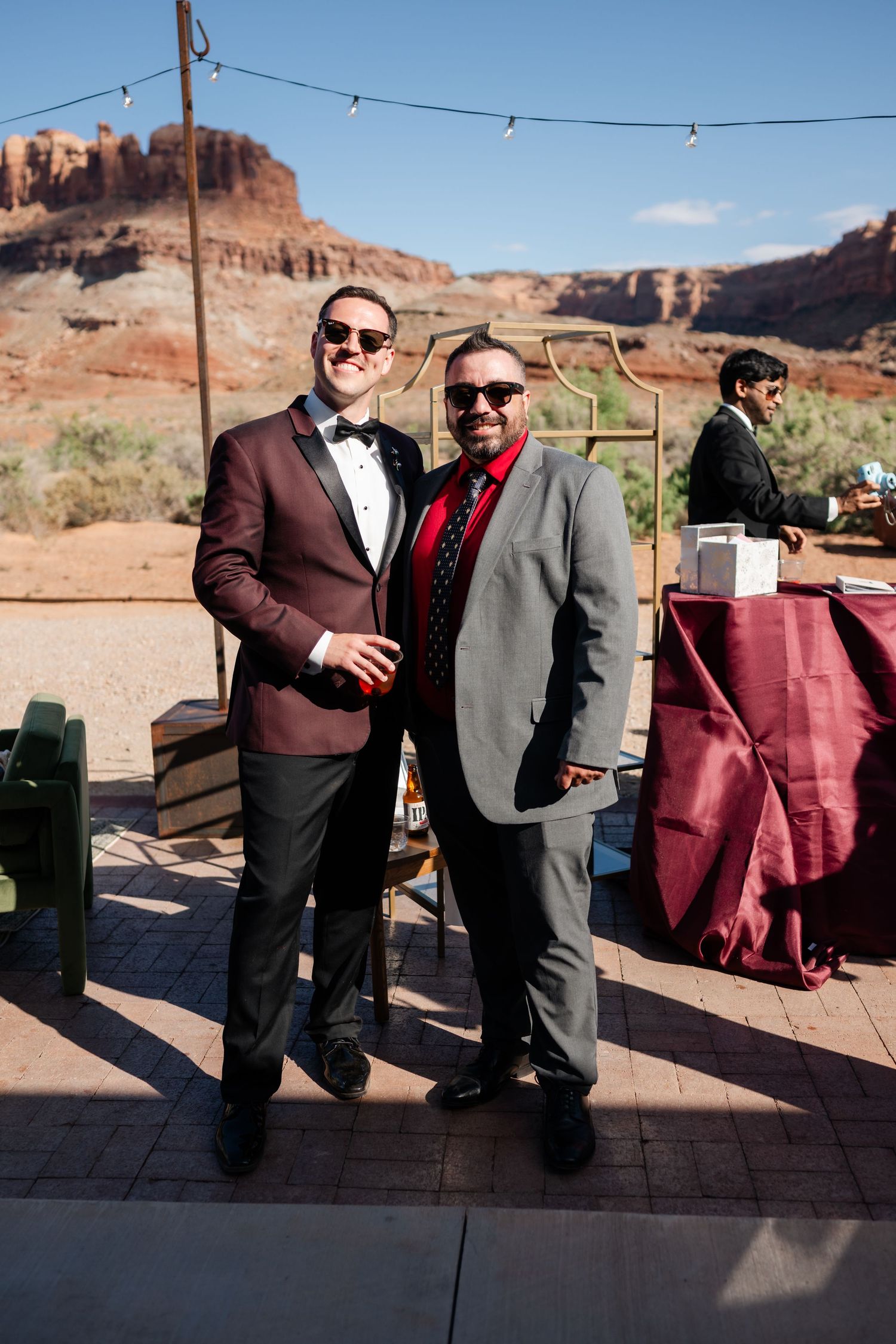 Two men in formal attire pose outdoors at a desert wedding venue with red rock formations in the background.