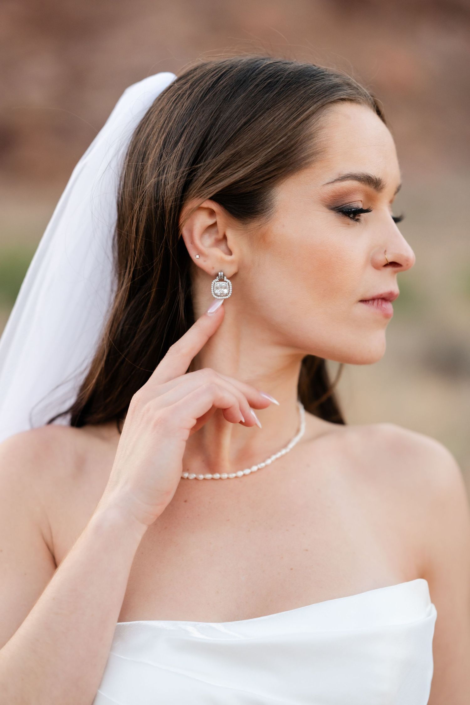 Bride in white strapless gown wearing pearl necklace and crystal drop earrings with veil in desert setting.