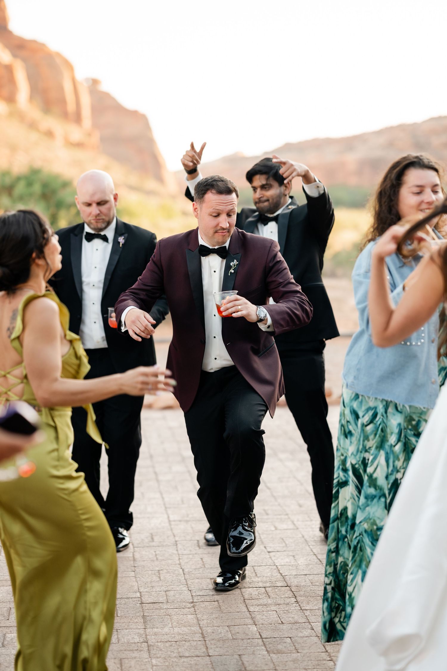 Groom in burgundy tuxedo dances with wedding guests outdoors against stunning red rock canyon backdrop.