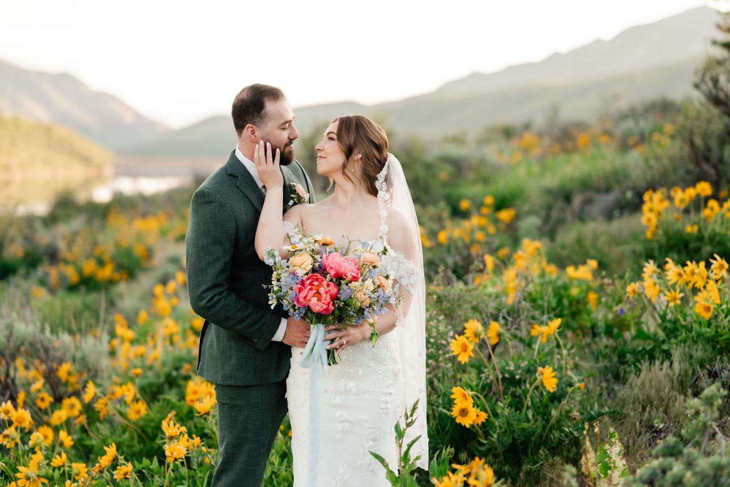A couple shares romantic moments among vibrant yellow wildflowers in a mountainous landscape at sunset.