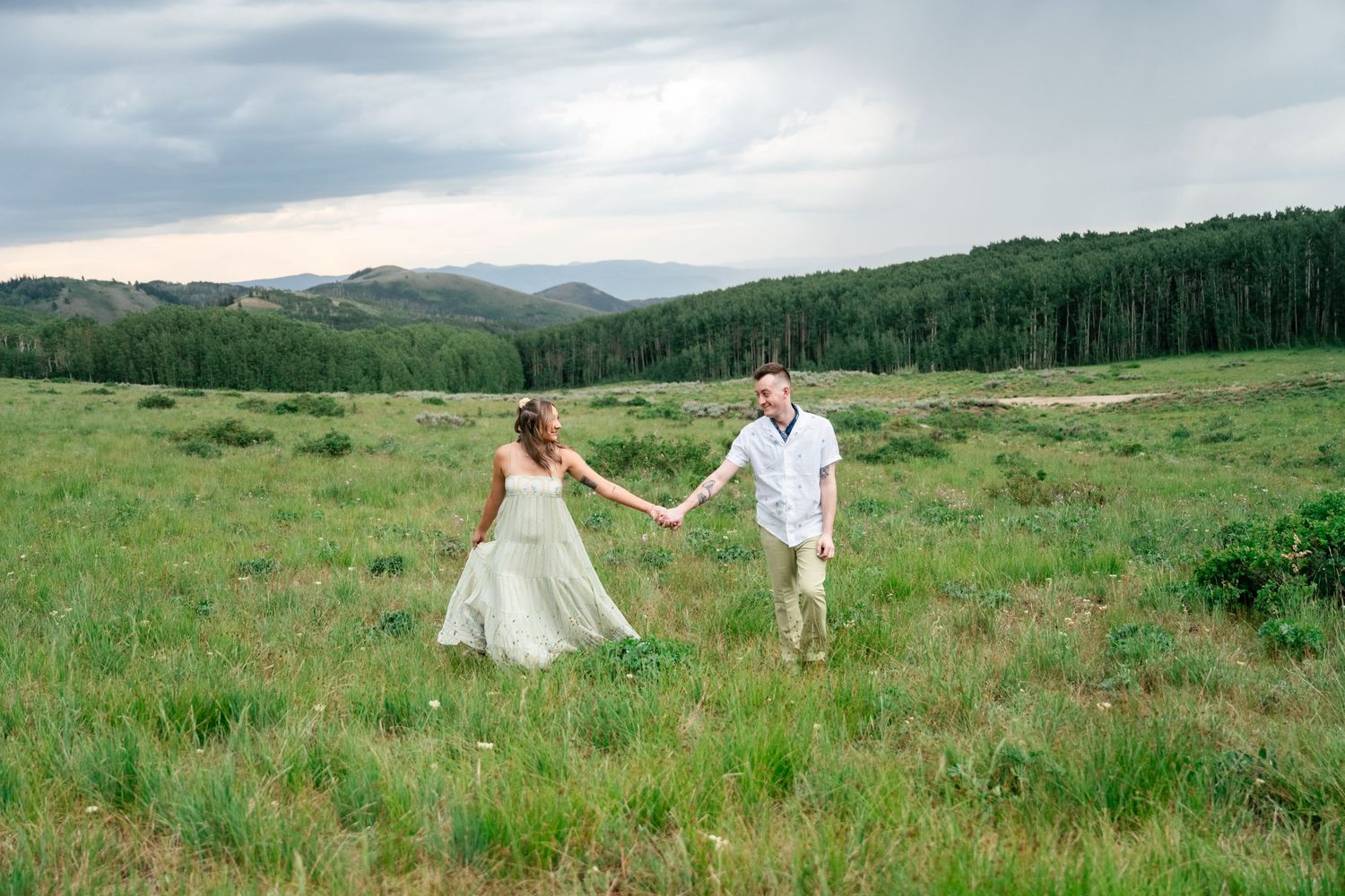A couple dances and twirls together in a scenic mountain meadow surrounded by green grass and rolling hills.