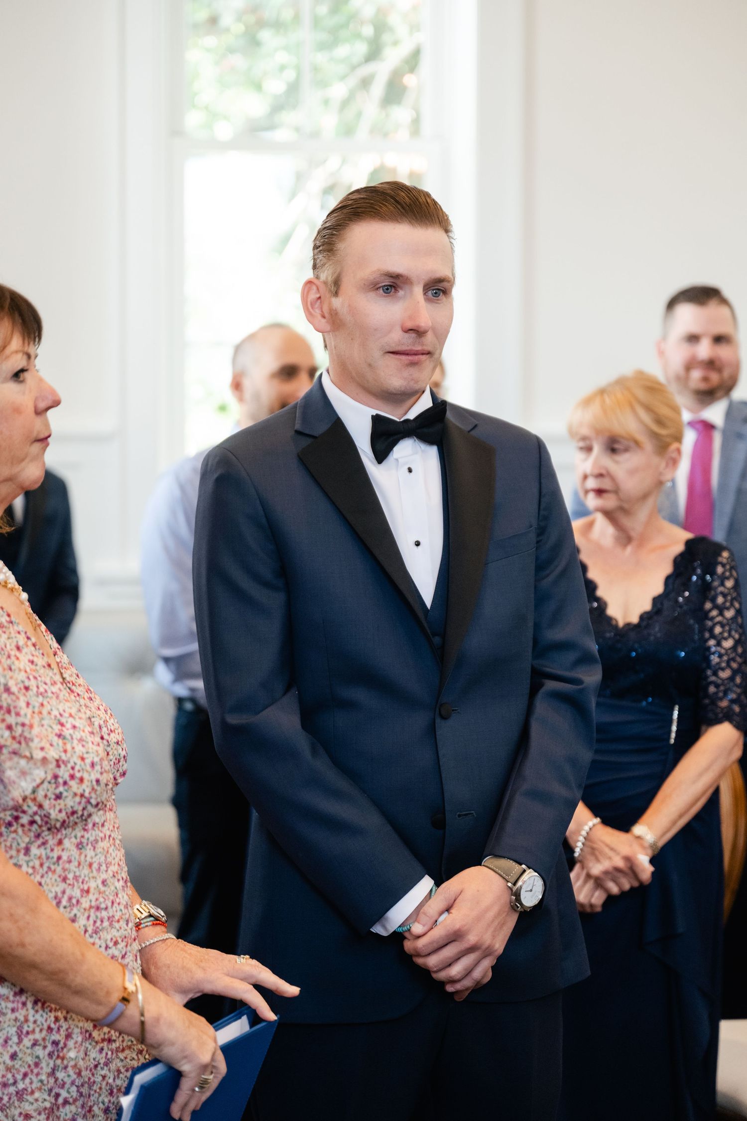 Wedding party members stand formally in a bright indoor venue wearing black tuxedos.