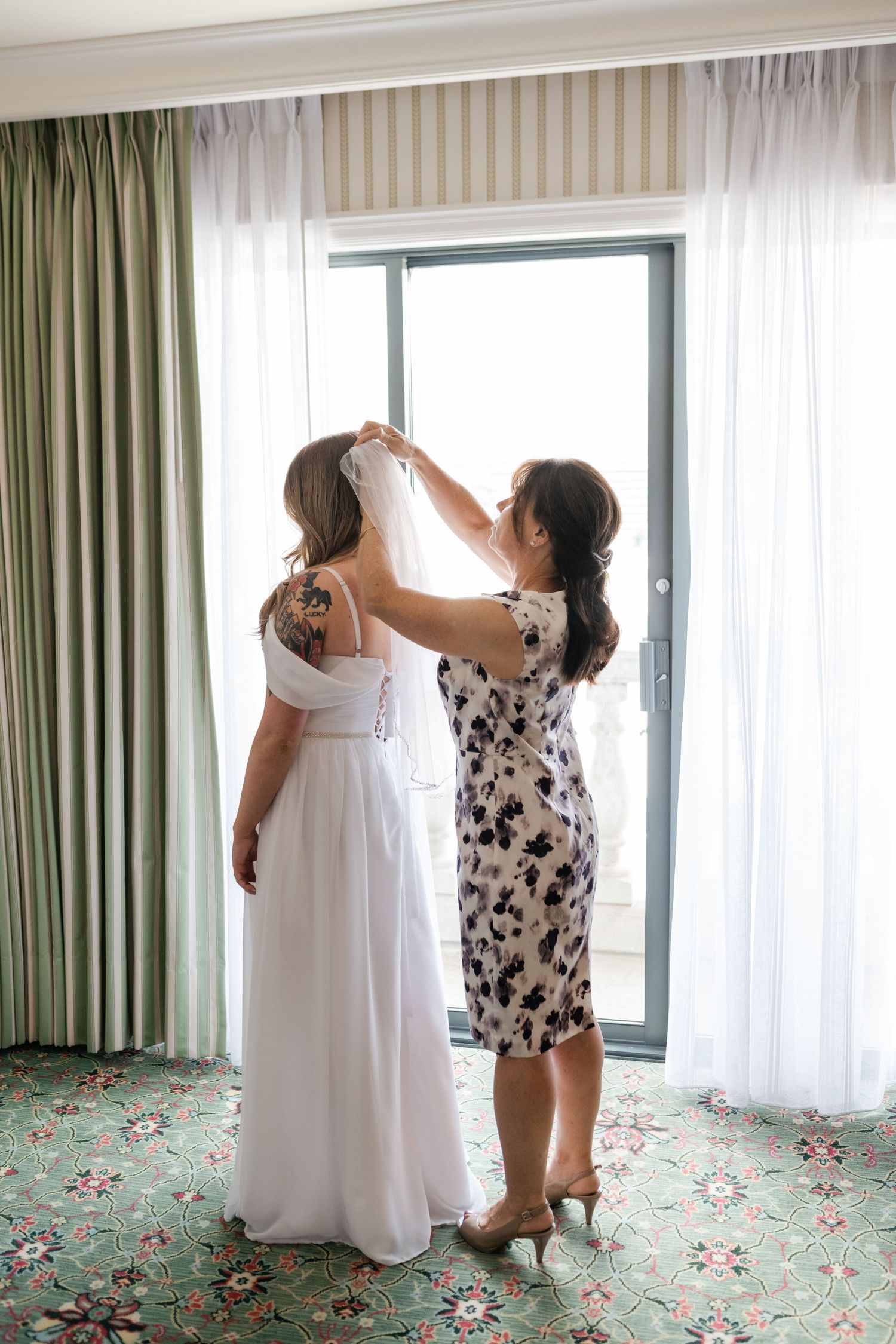 Two people in white and floral dresses adjust curtains in a hotel room with green carpet.