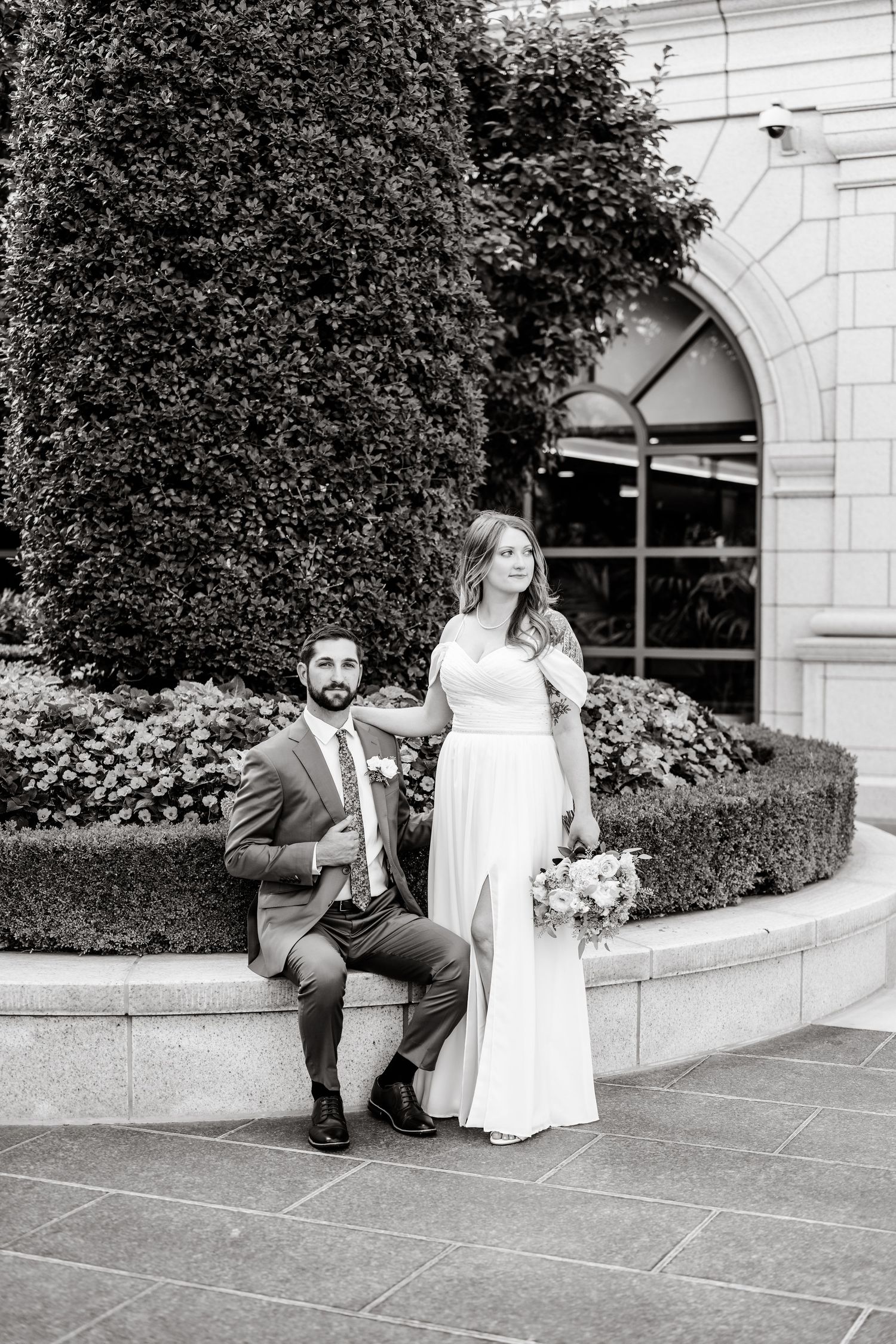 A wedding couple sits on stone steps beside manicured hedges at an elegant building entrance.