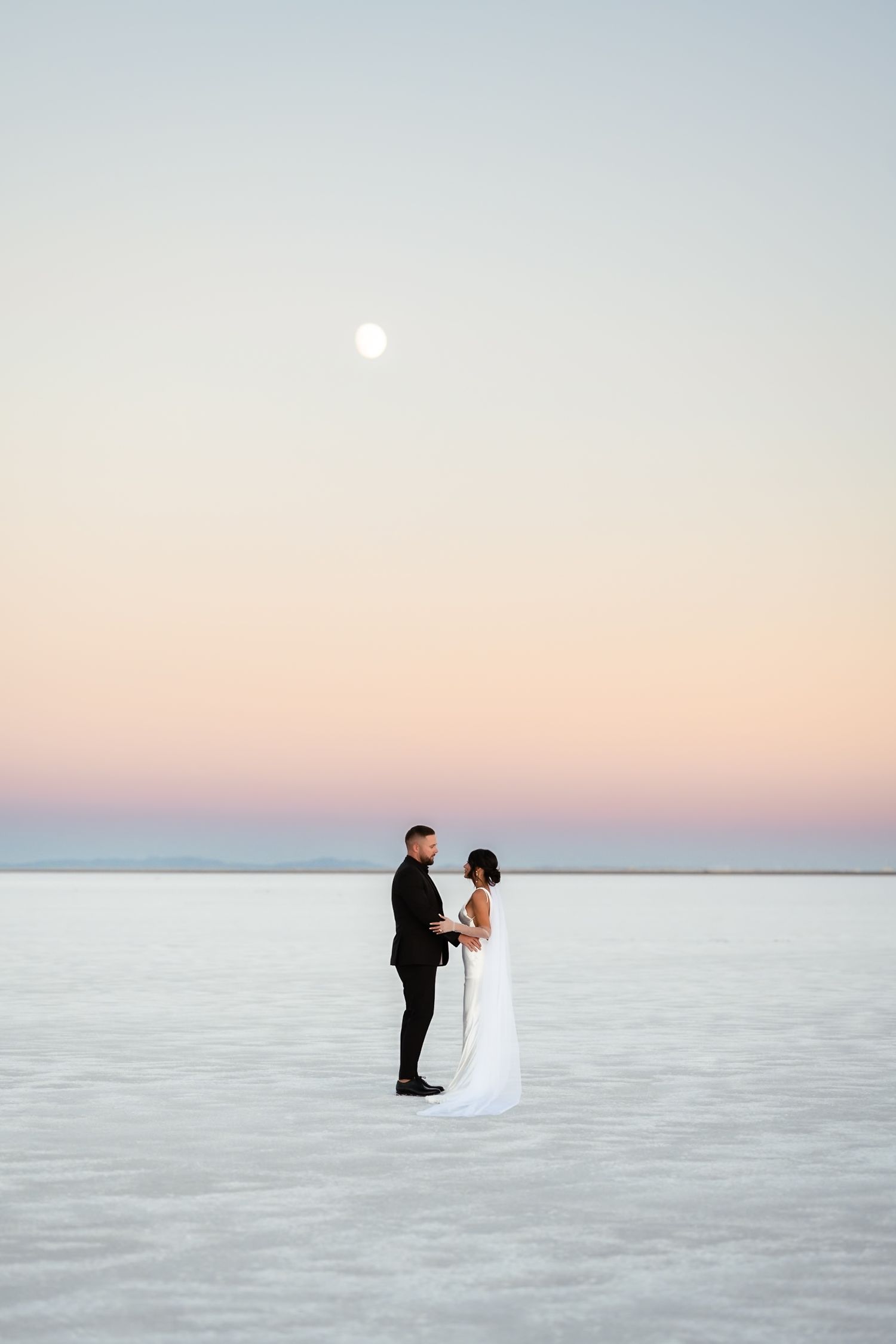 A romantic wedding photo of a couple standing on salt flats under a full moon at dusk.