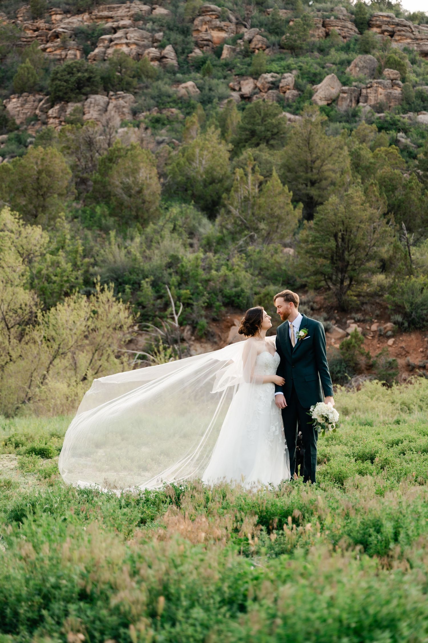 Wedding couple poses in front of red rock formations as a long veil flows in the wind.