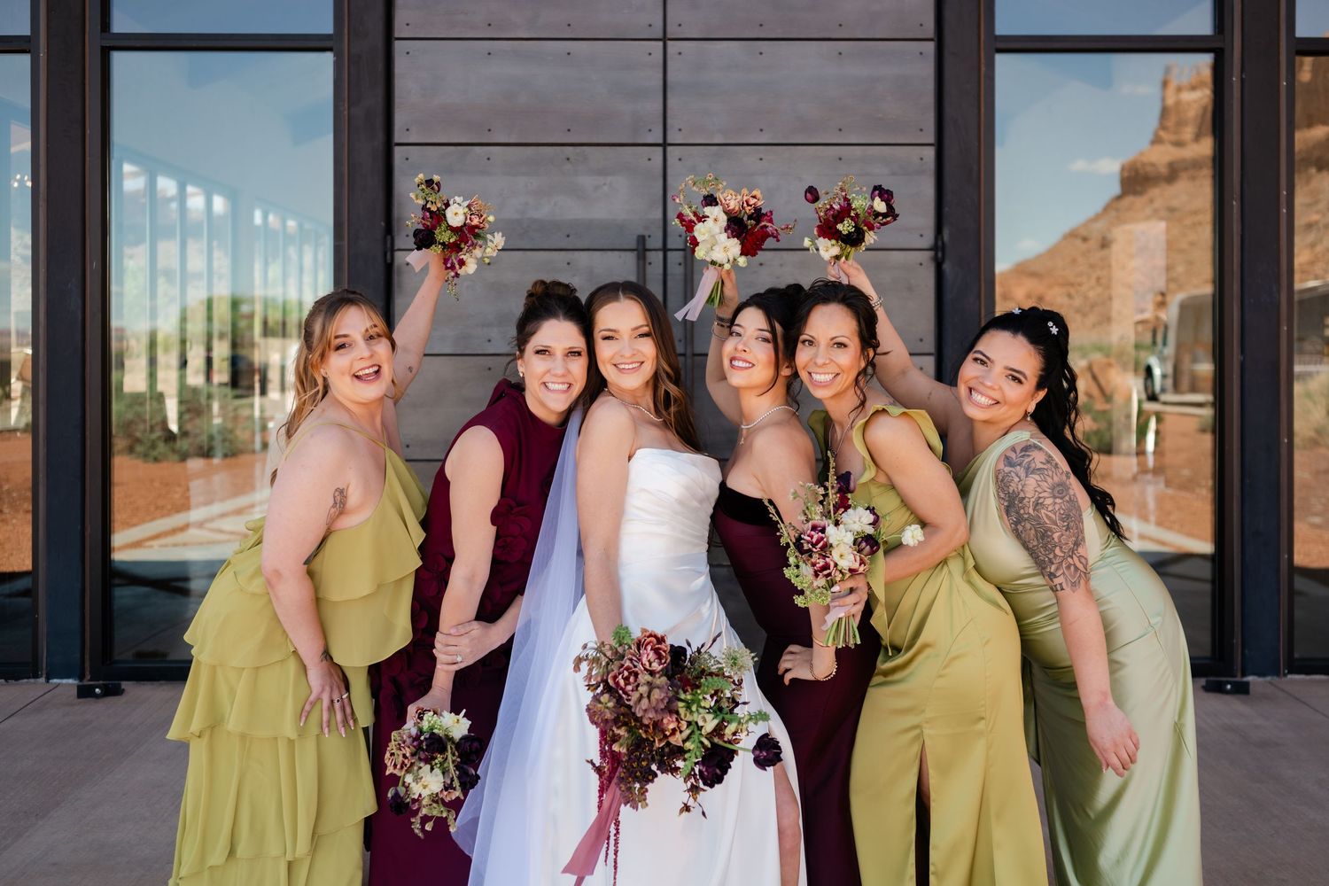 Bride in white gown poses with bridesmaids in burgundy and olive green dresses holding bouquets outside modern venue.