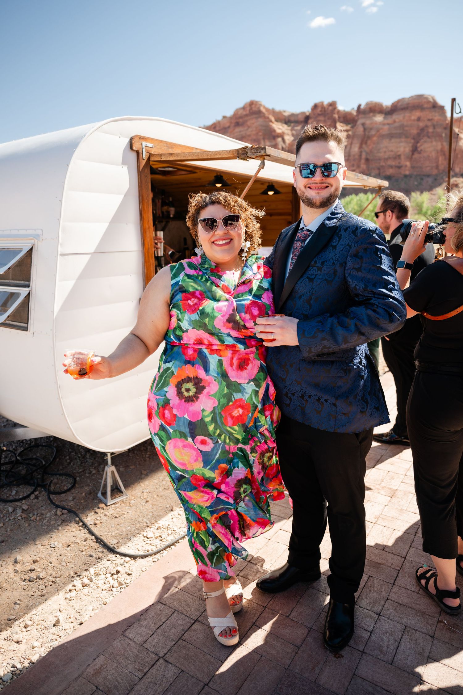 Couple in floral dress and navy suit posing by white vintage trailer with red rock formations backdrop.