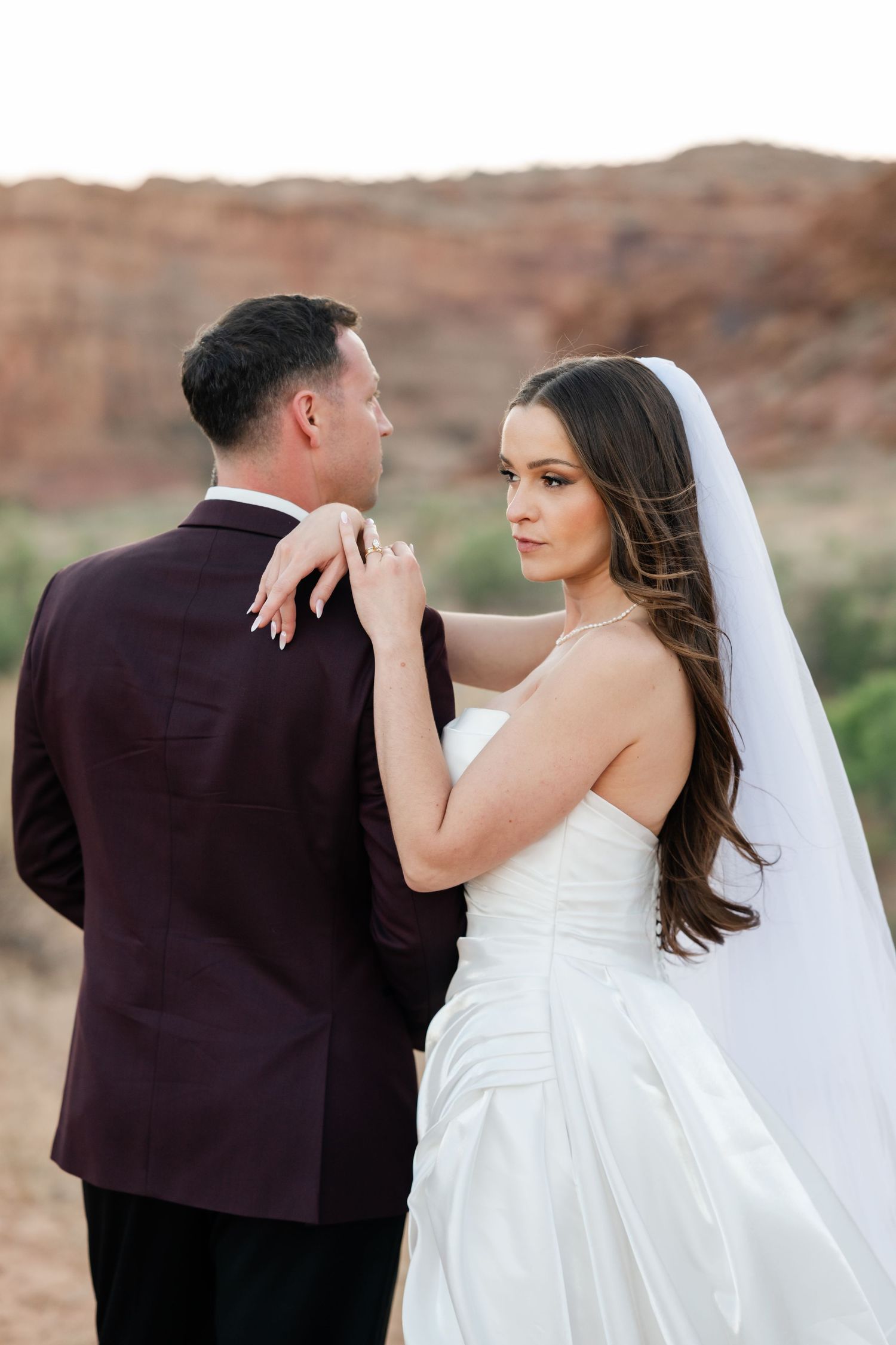Bride in strapless white gown looks over shoulder while resting hands on groom in burgundy tuxedo, red rocks behind them.