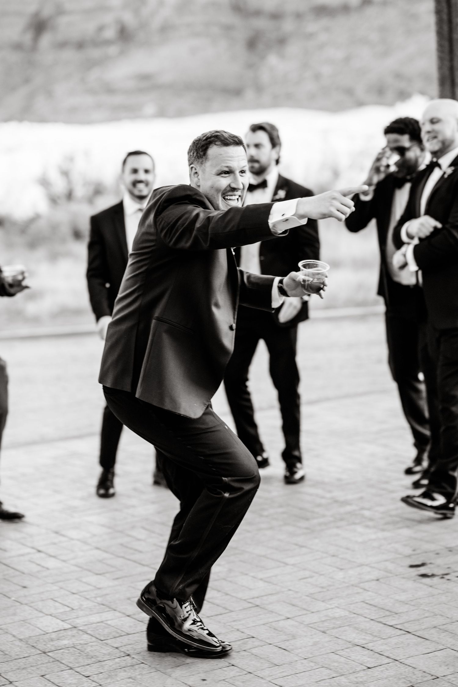 Man in tuxedo dancing and pointing at wedding reception outdoors in black and white photo.
